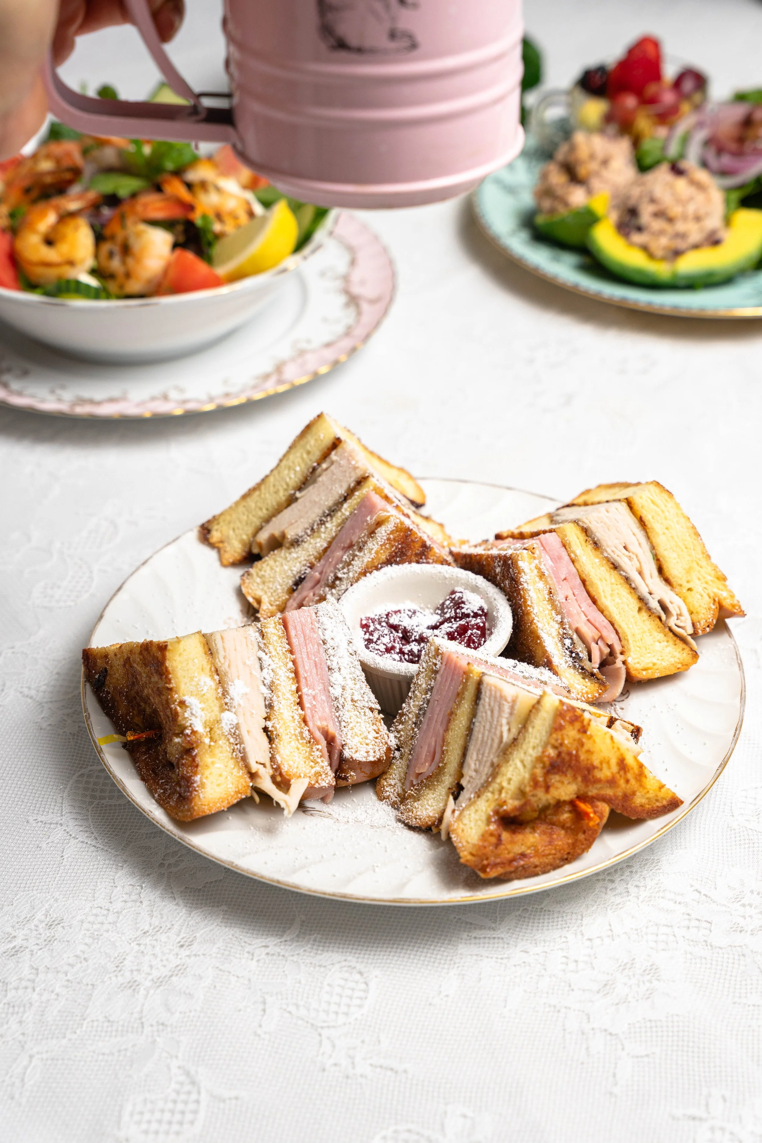 Plate of sliced Monte Cristo sandwiches with powdered sugar and raspberry jam, shrimp salad in the background, and pink pitcher being poured.