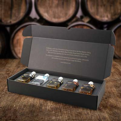 A black box containing five miniature bottles of whisky, each with a different colored label and sealed with a white cap, placed on a wooden surface with barrels in the background.