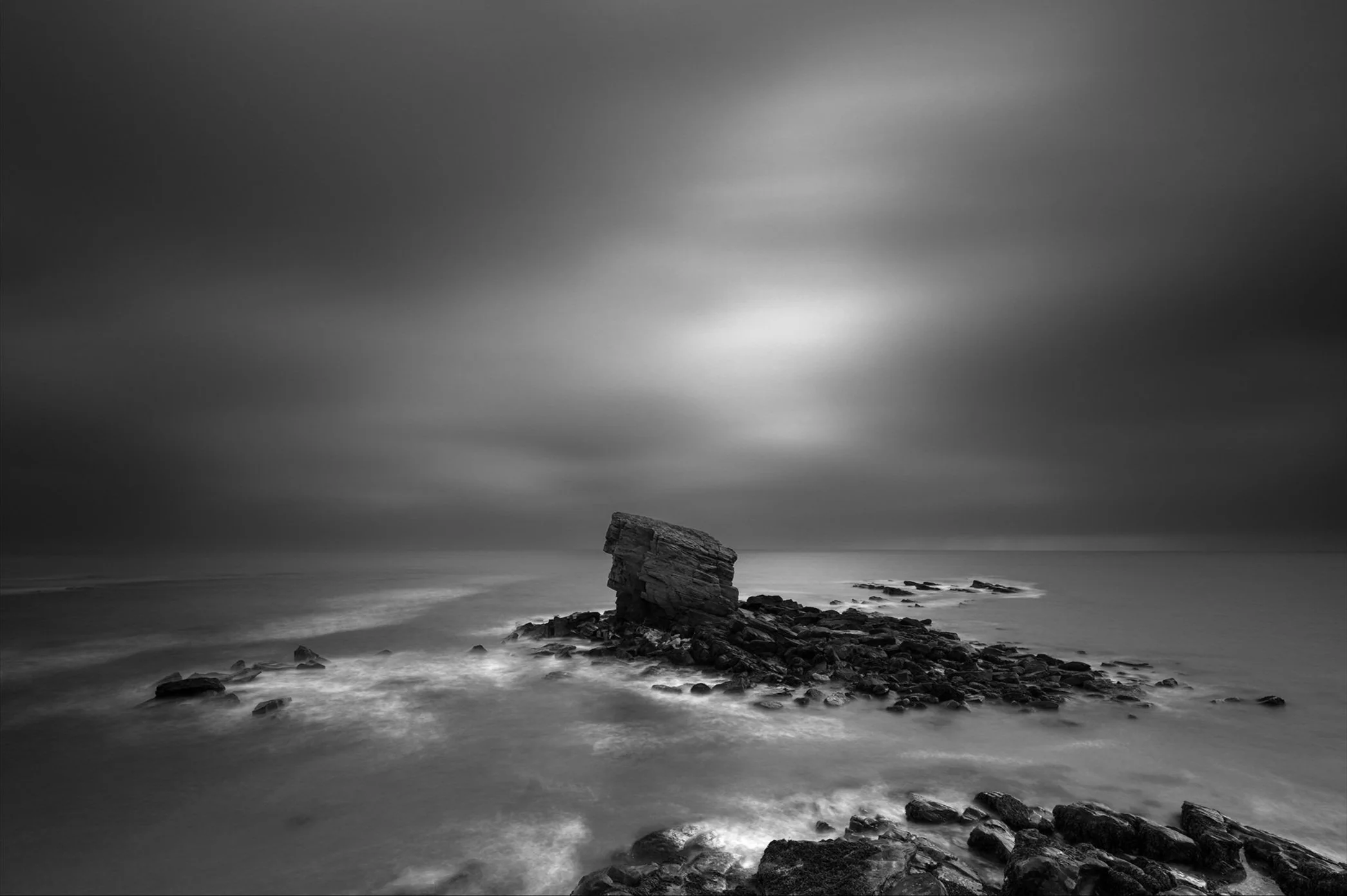 Black and white photograph of a rocky seashore with a large, tilted rock formation standing isolated on smaller rocks, under a cloudy sky.