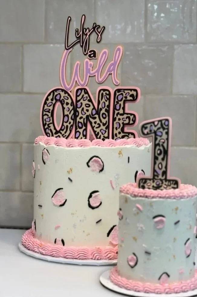 Two celebration cakes decorated with pink and black speckles, with pink icing borders. One large cake has a topper reading "Leigh's a Wild ONE" and the smaller cake has a large number one shaped topper, all with matching leopard print patterns.