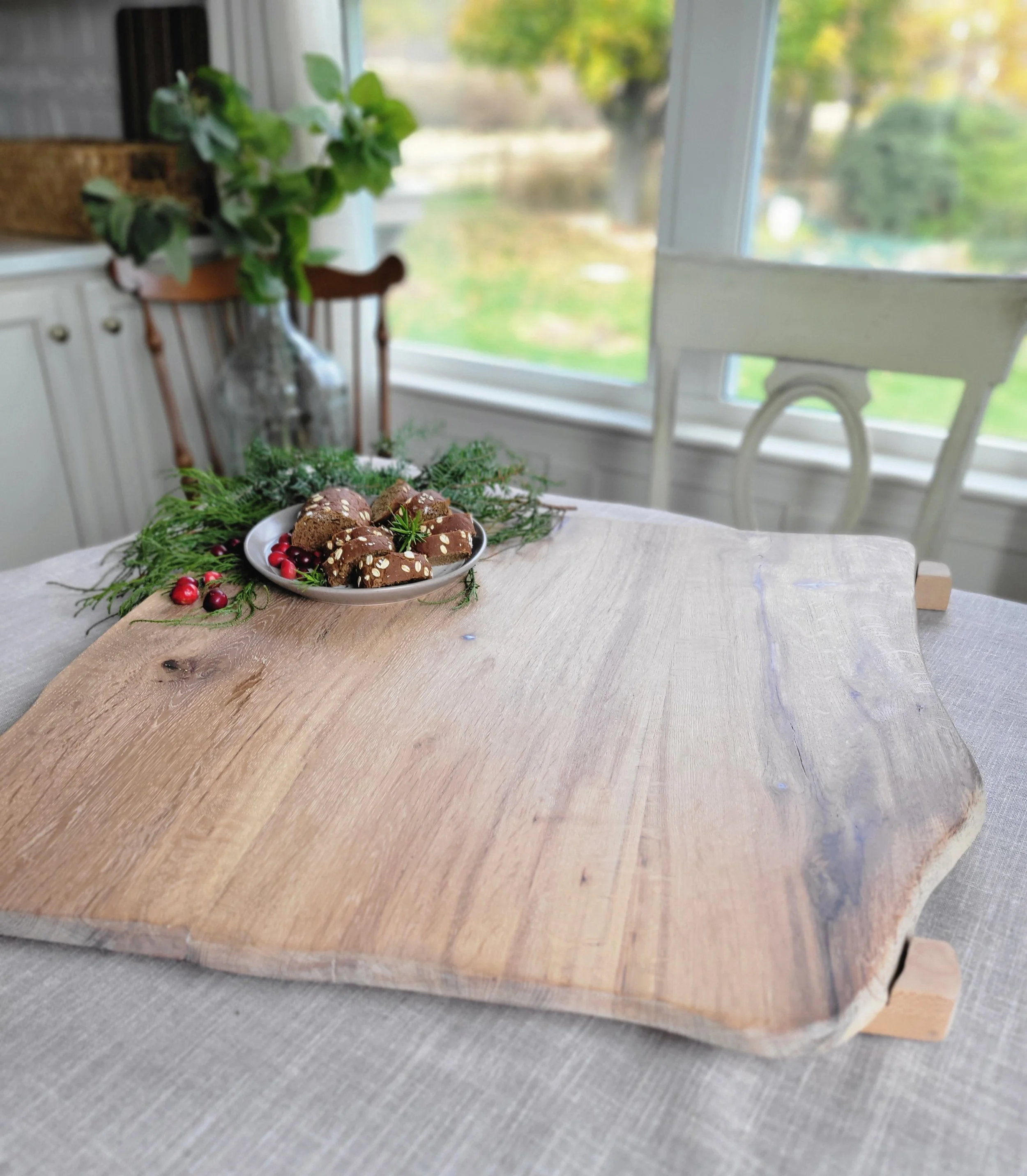 A wooden serving board on a table with a plate of cookies decorated with white sprinkles and red berries, with a sprig of greenery, near a window with a blurry view of the outdoors.