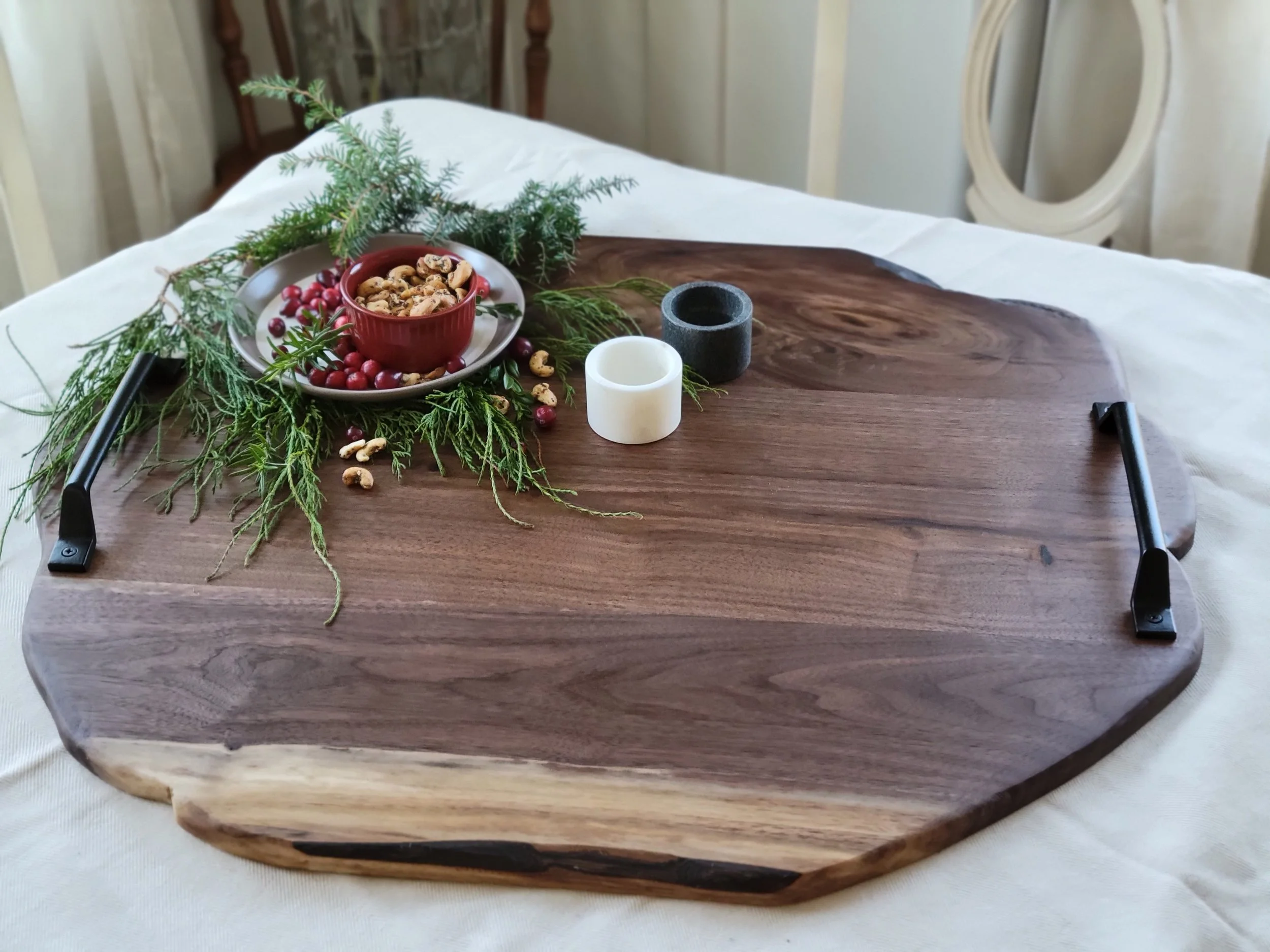 Decorative wooden serving tray with greenery, a small bowl of mixed nuts, two candle holders, and cranberries on a table.