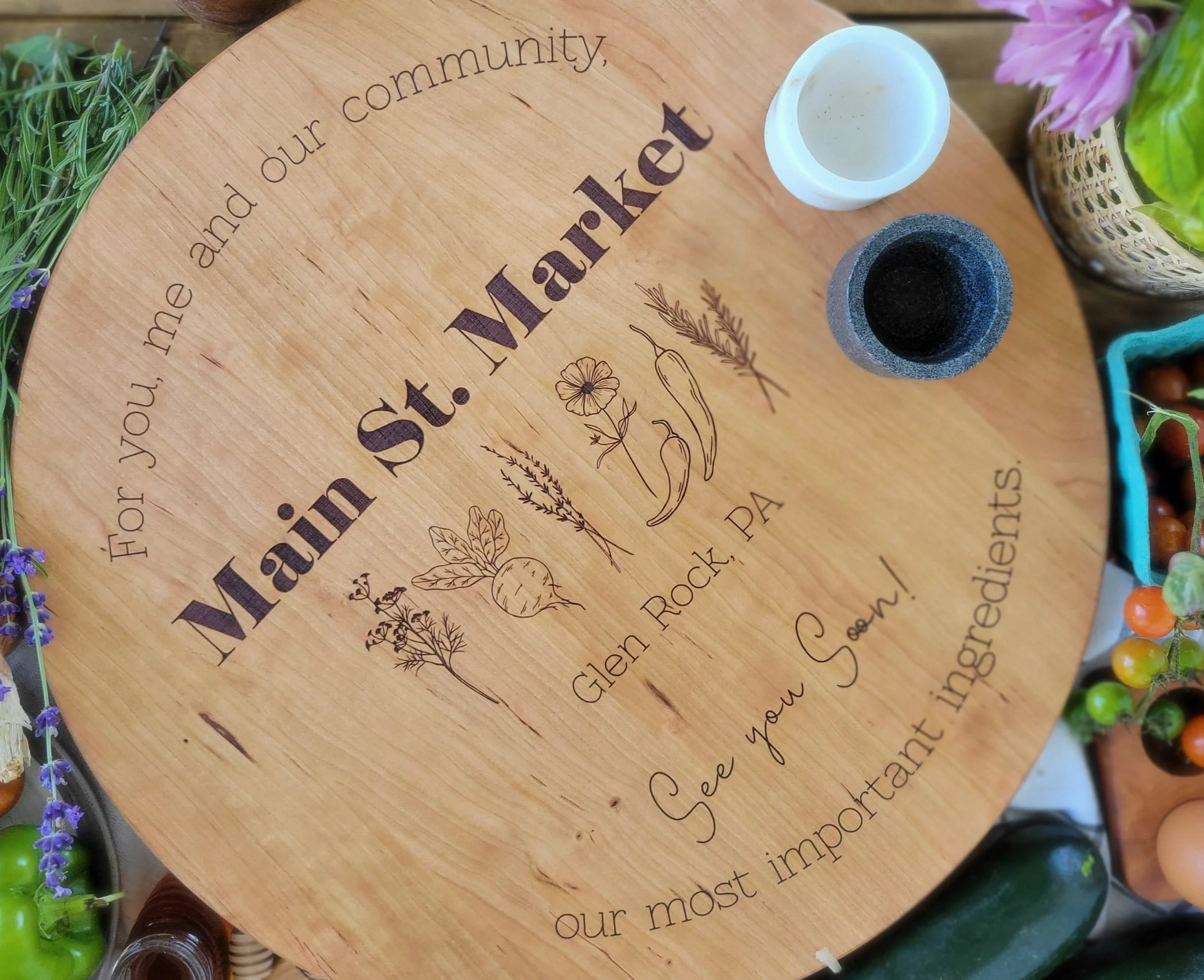 A round wooden cutting board with engraved text and illustrations, surrounded by various plants and vegetables, including a zucchini, berries, and flowers.