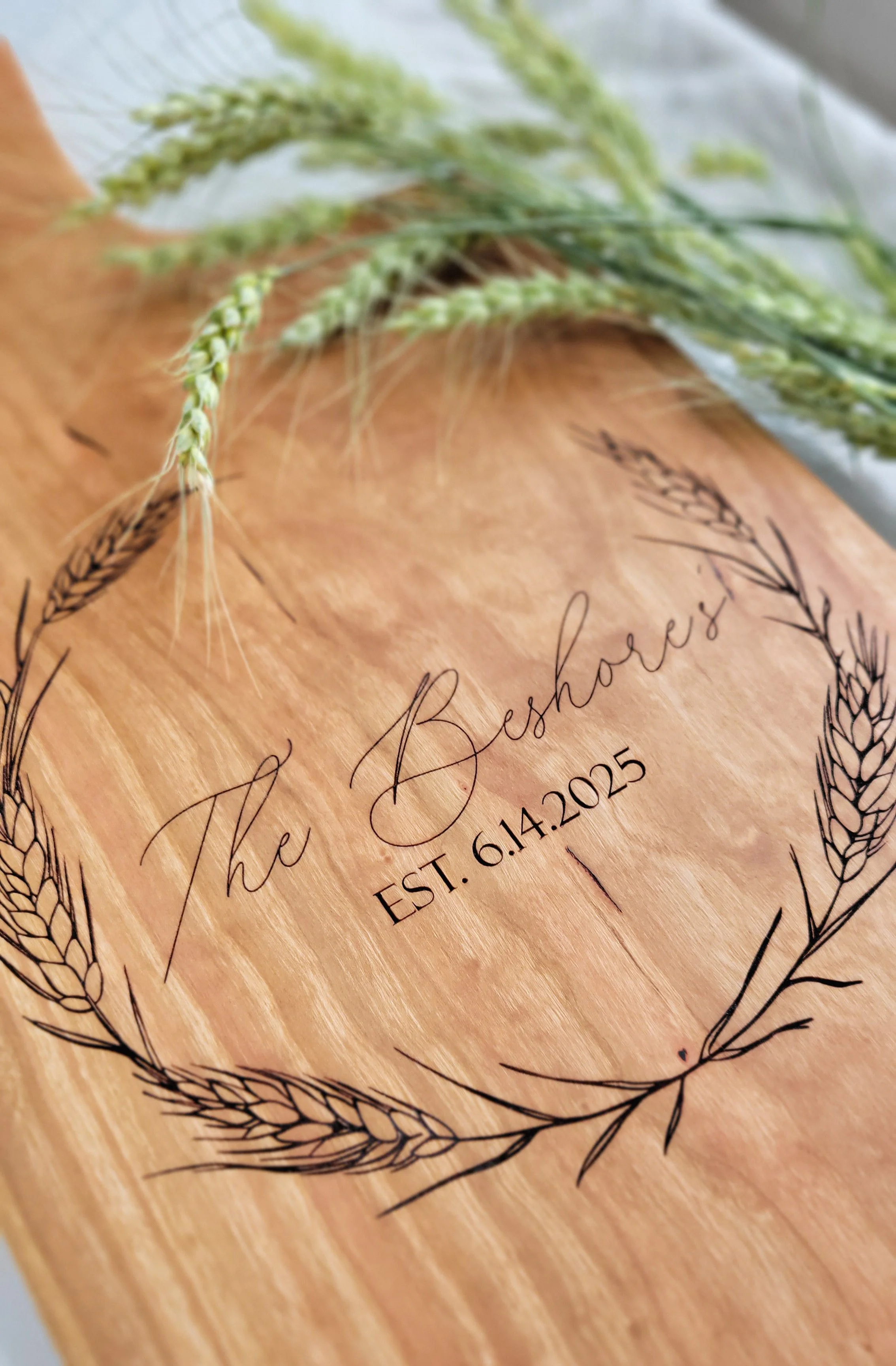 A wooden plaque with engraved text that reads "The Beshore's EST. 6.14.2015" inside a decorative wheat wheat circle, with some green wheat stalks resting on top.