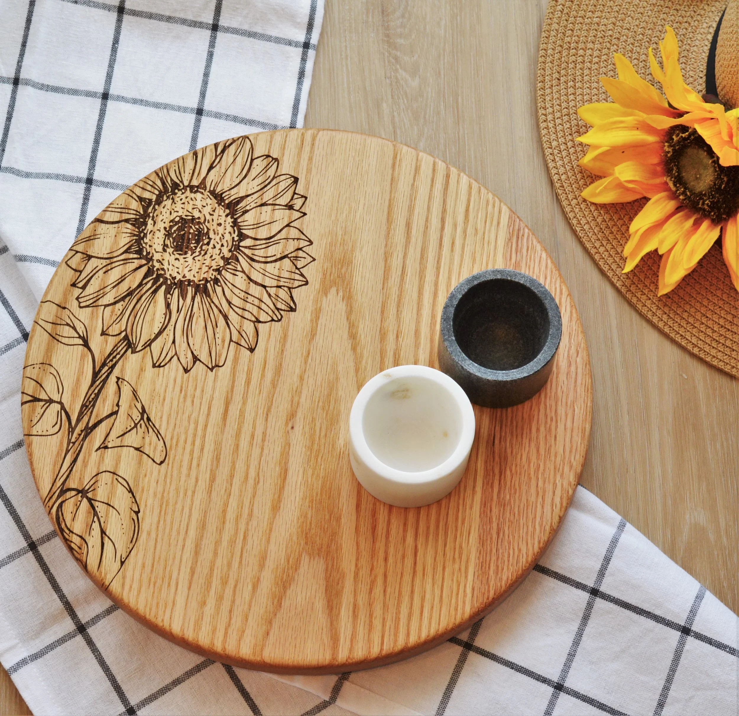 Decorative wooden serving board with sunflower illustration, two small containers, and a sunflower on a woven placemat, with a checked cloth underneath.
