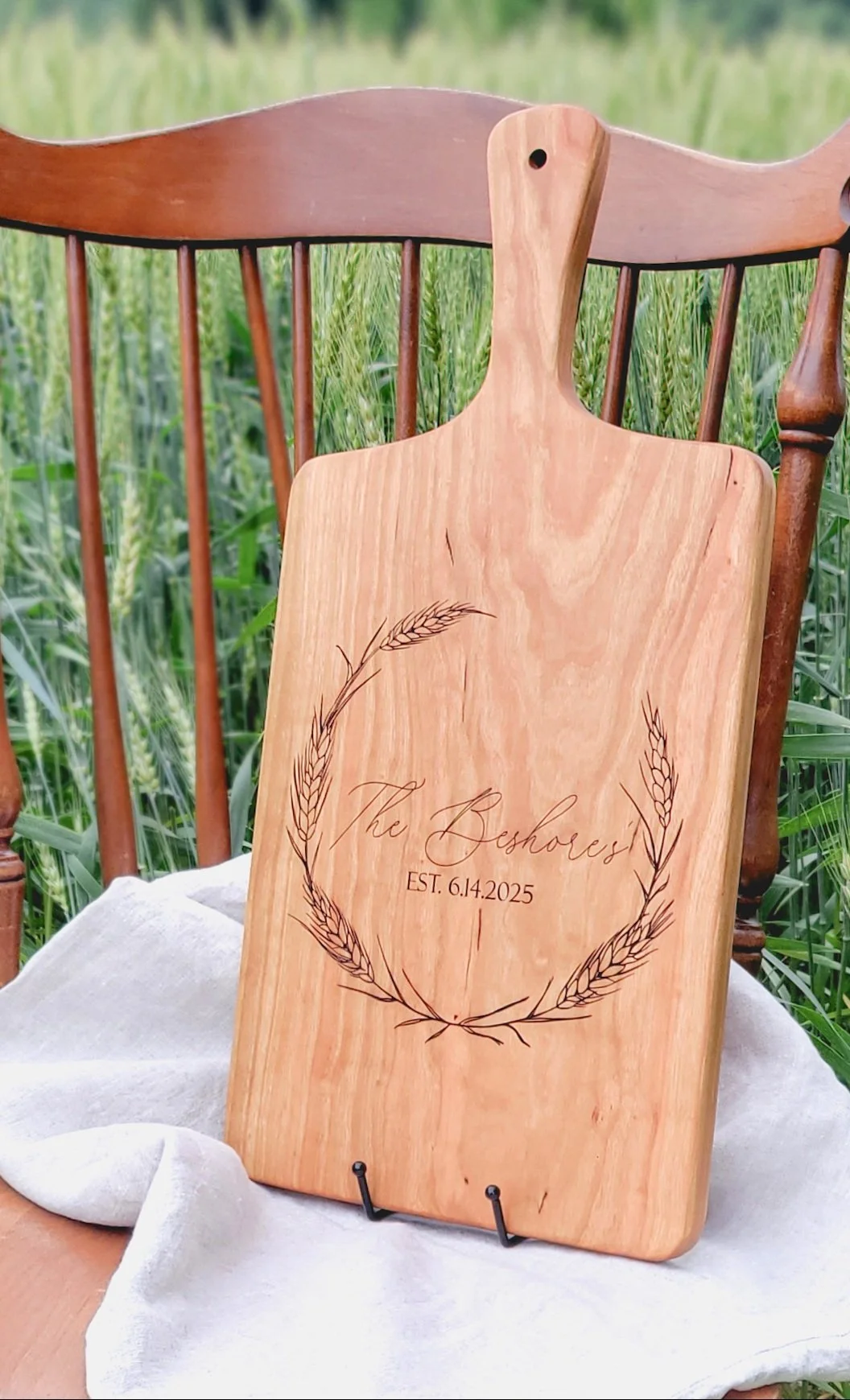 A wooden sign engraved with the words "The Beshores" and the date "EST. 6.14.2025" within a wheat wreath design, placed on a stand outdoors in a grassy field.