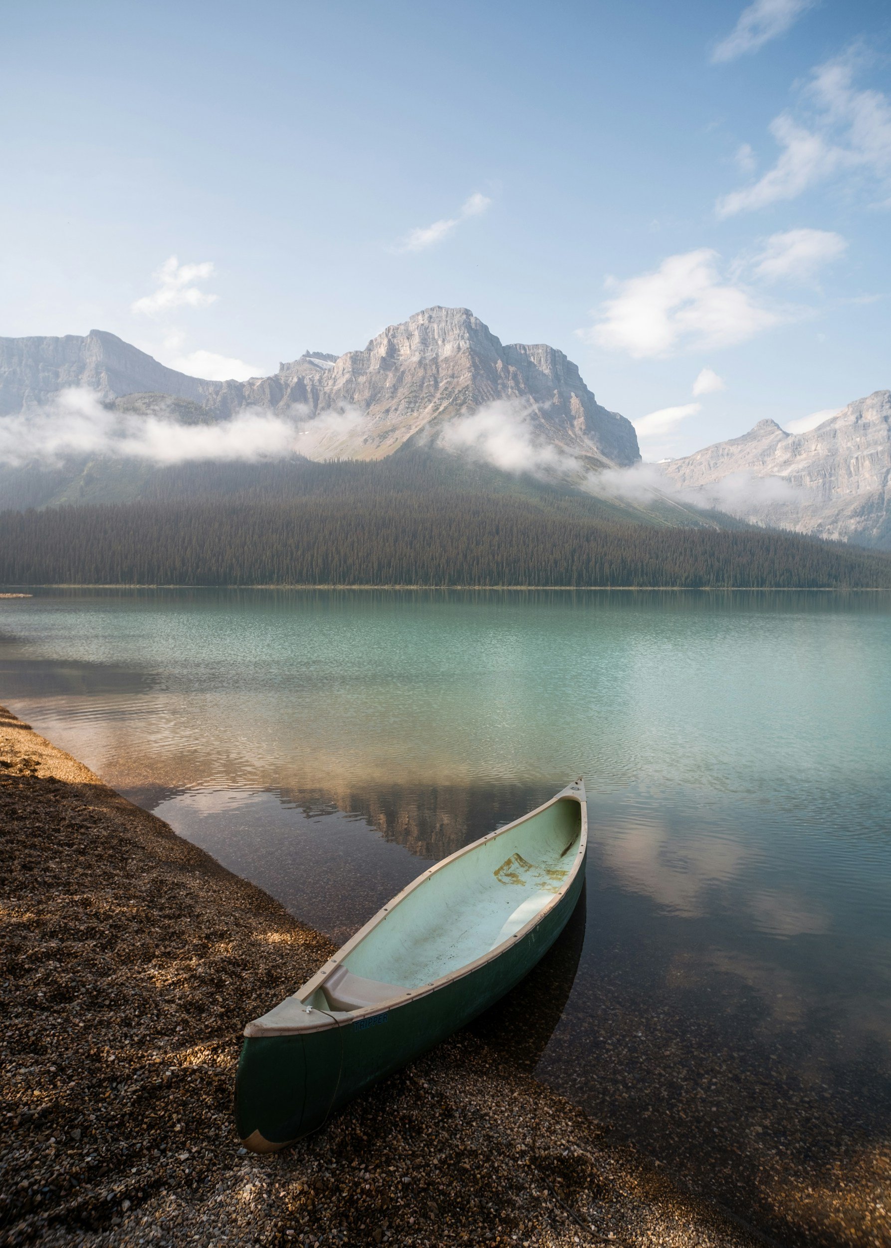 A boat resting on the edge of a lake with calm water, mountainous landscape with lush forest in the background, and partly cloudy sky.
