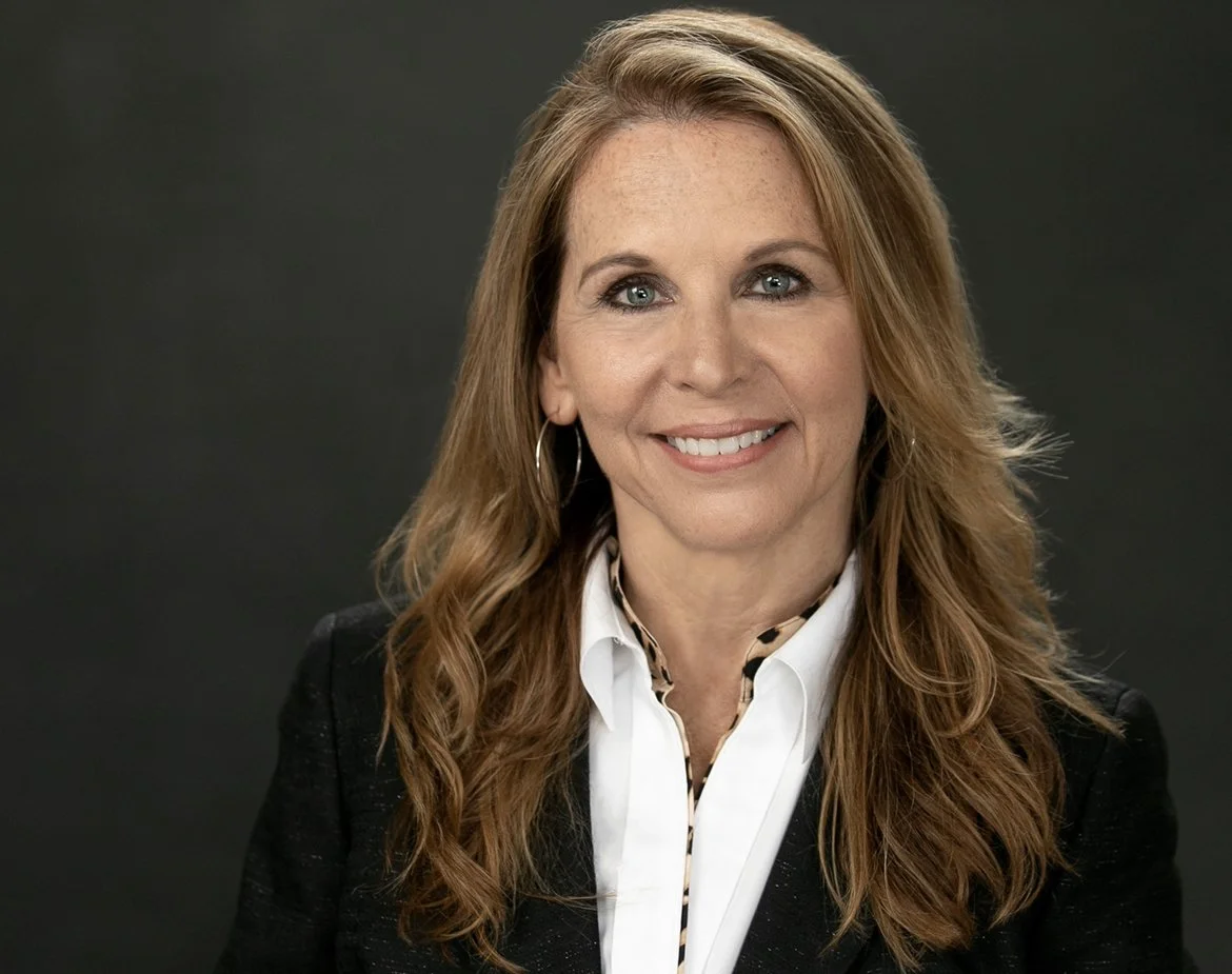 Dr. Jacquelyn Paykel with long, wavy reddish hair, blue eyes, and a bright smile, wearing a black blazer and white shirt, posing against a dark background.