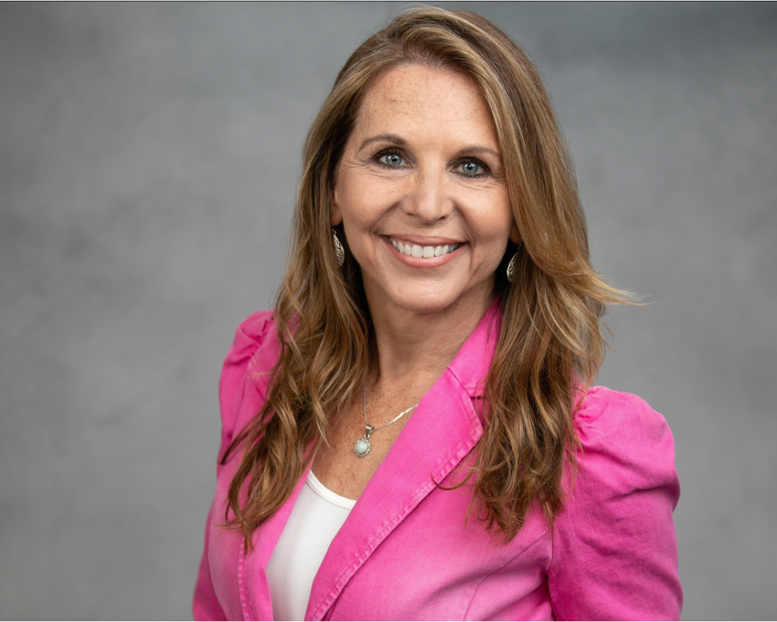 Portrait of Dr. Jacquelyn Paykel wearing a pink blazer, a white top, and jewelry, smiling against a gray background.