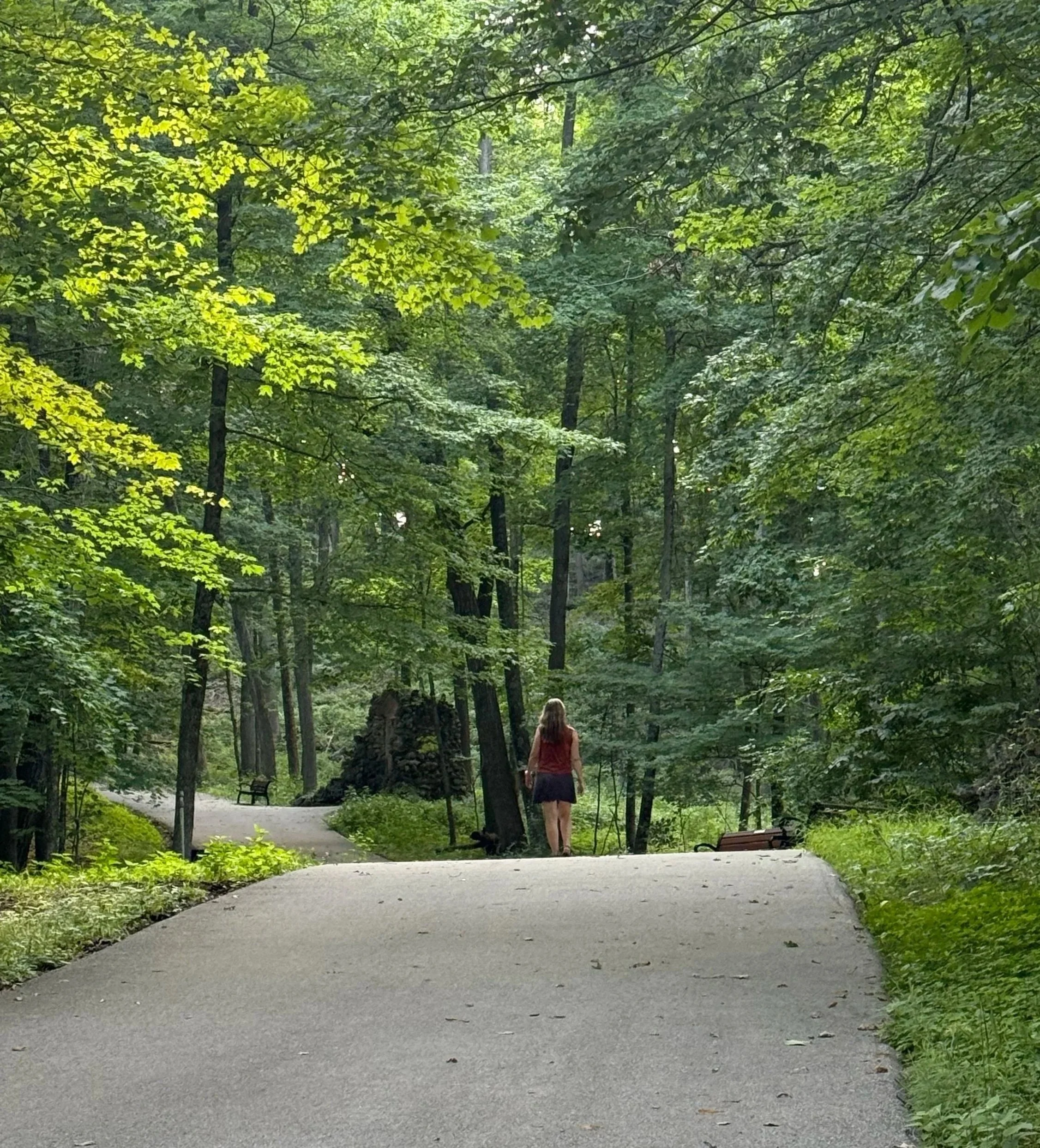 A woman walking on a paved trail through a lush green forest with tall trees and dense foliage.