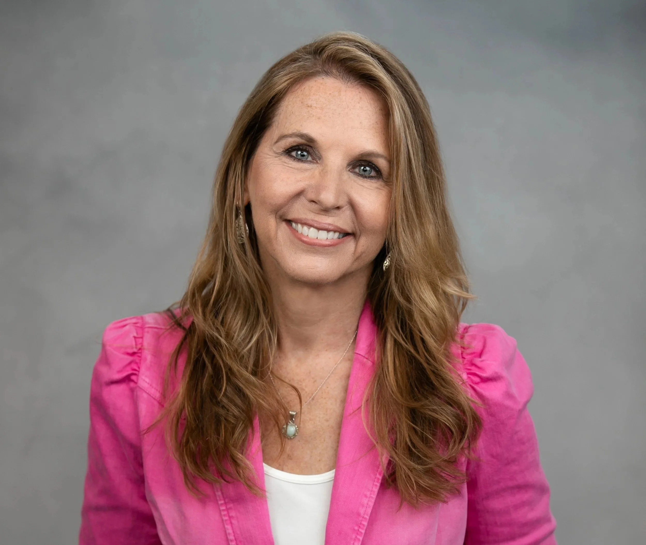 Dr. Jacquelyn Paykel, reddish-brown hair, blue eyes, and a friendly smile, wearing a pink blazer over a white top, with a pearl necklace and earrings, posed against a gray background.