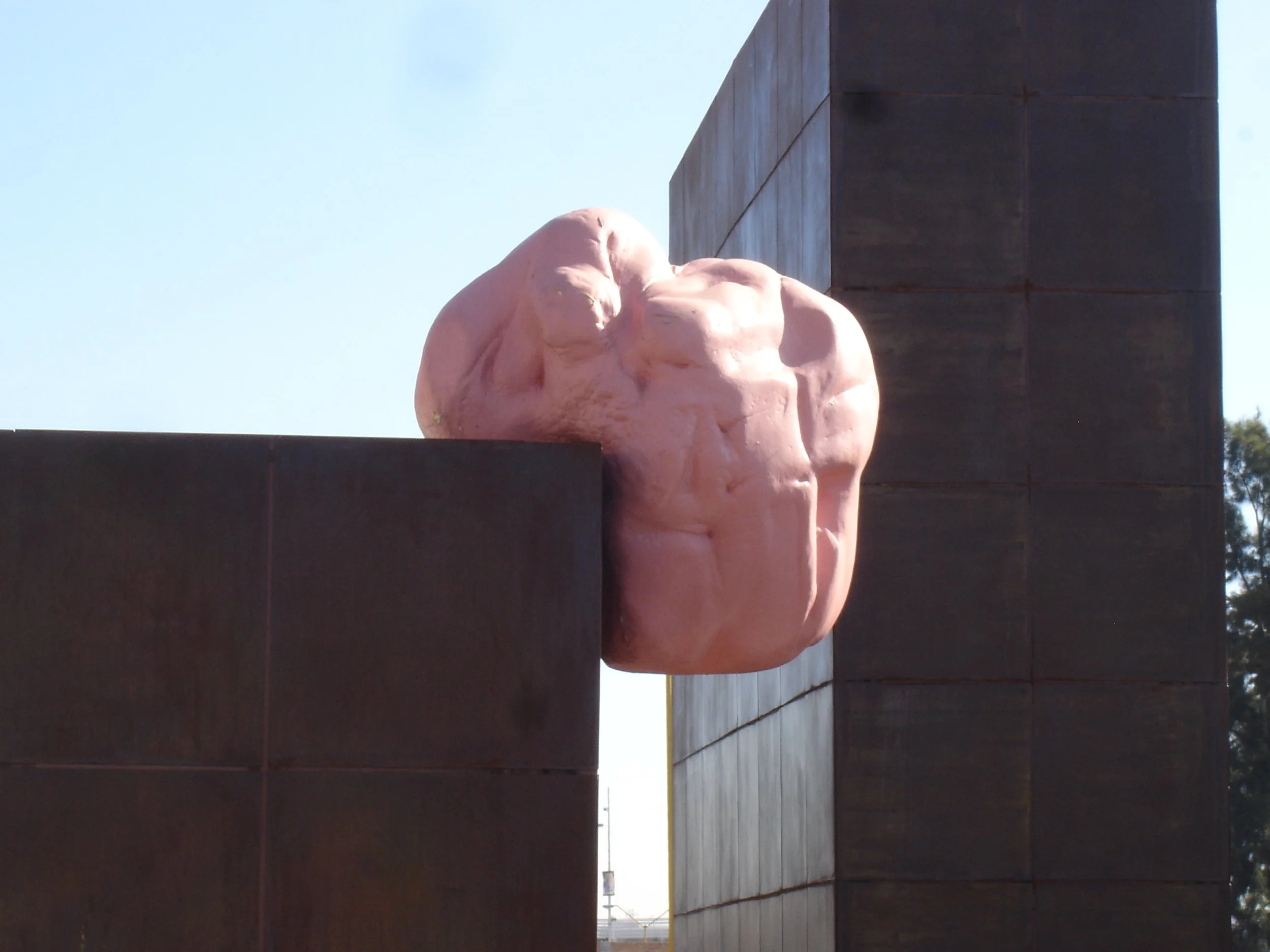 Image of an outdoor sculpture of a massive pink piece of chewing gum resting on a big dark brown structure with a clear sky background. this piece is itinerant and photographed in different spot in Aguascalientes City.