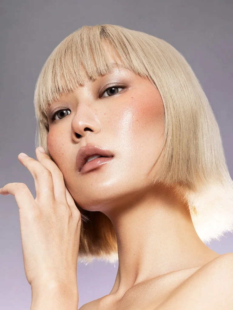 Close-up portrait of a woman with short blonde hair, natural makeup, and fair skin posing with her hand near her face against a plain gray background. Beauty Photographer Los Angeles.