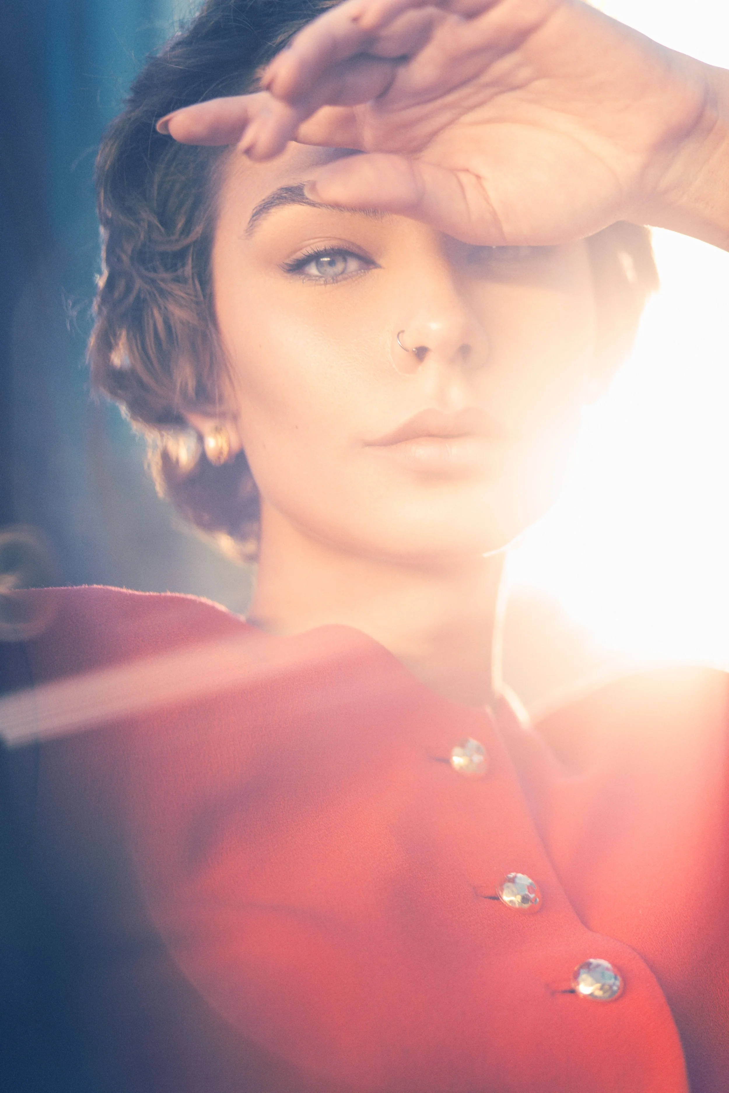 A woman with short wavy hair, light skin, blonde eyebrows, wearing a nose ring, earrings, a red top with crystal buttons, and posing with her hand above her eyes in front of bright sunlight.