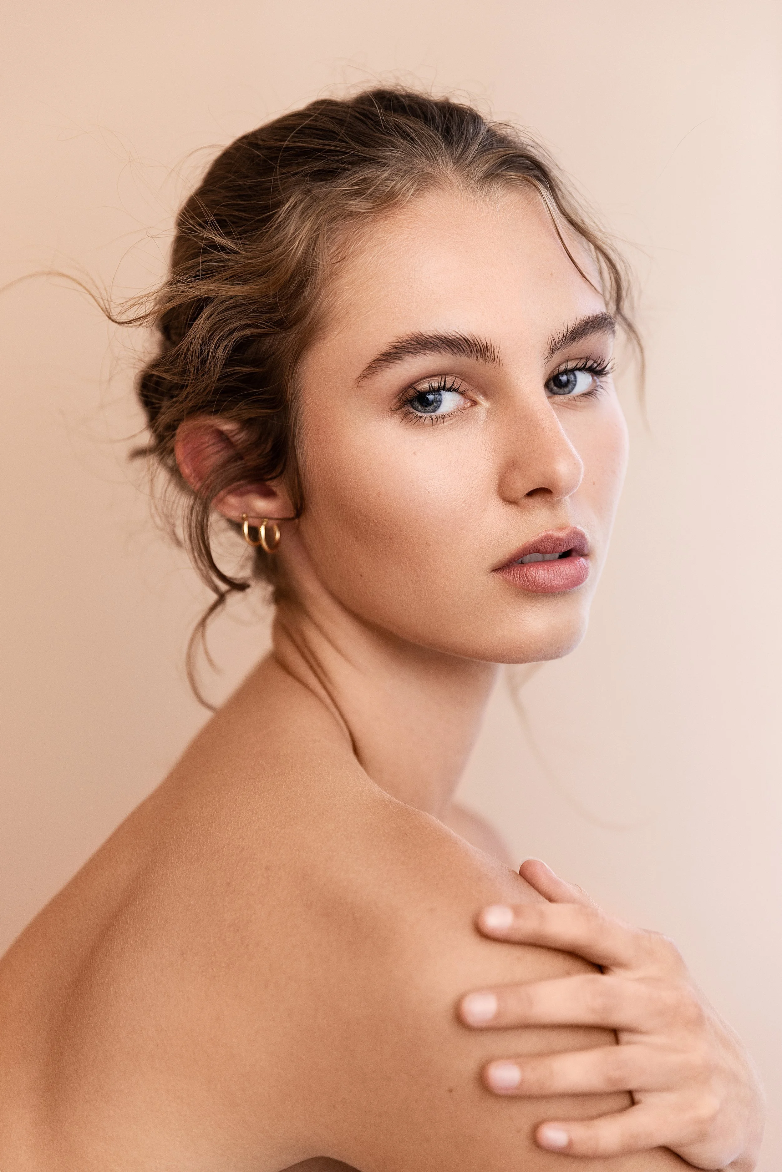 A portrait of a young woman with blue eyes, light brown hair styled in loose waves, wearing small gold hoop earrings, gently touching her shoulder, with a neutral beige background.