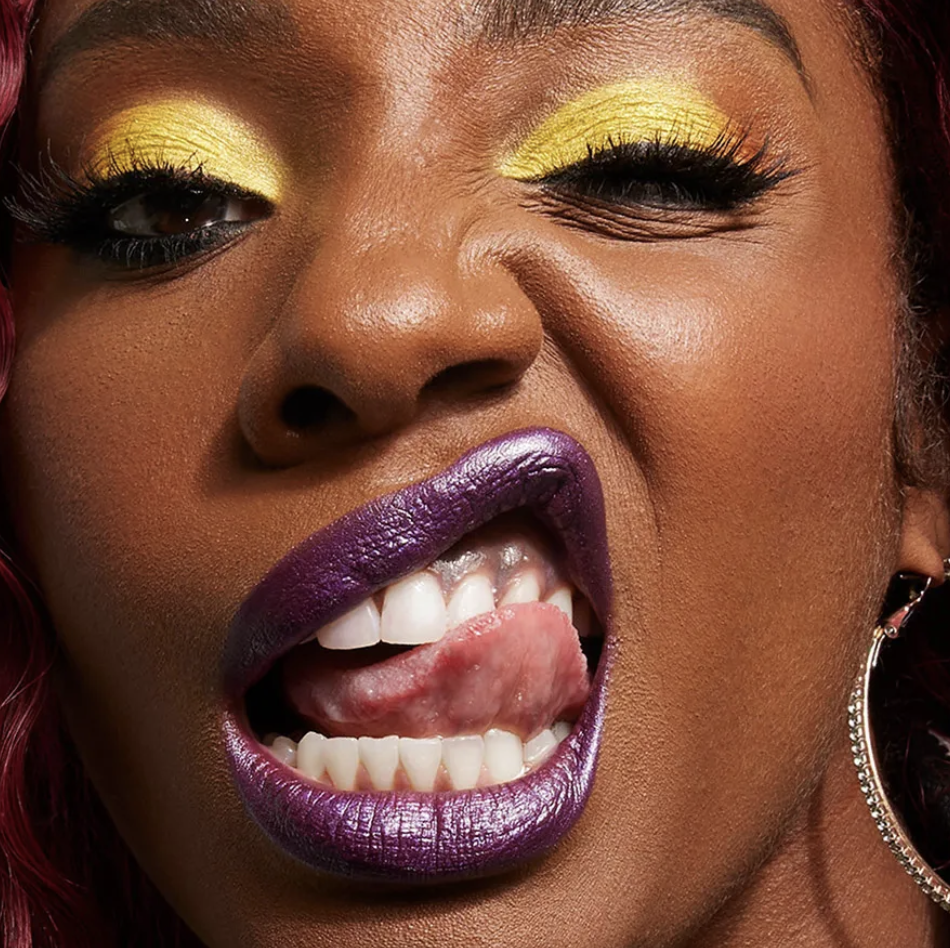 A close-up of a woman with bold makeup, wearing yellow eyeshadow and purple lipstick, sticking out her tongue with a roughened surface and white teeth visible. Commercial Photographer Los Angeles.