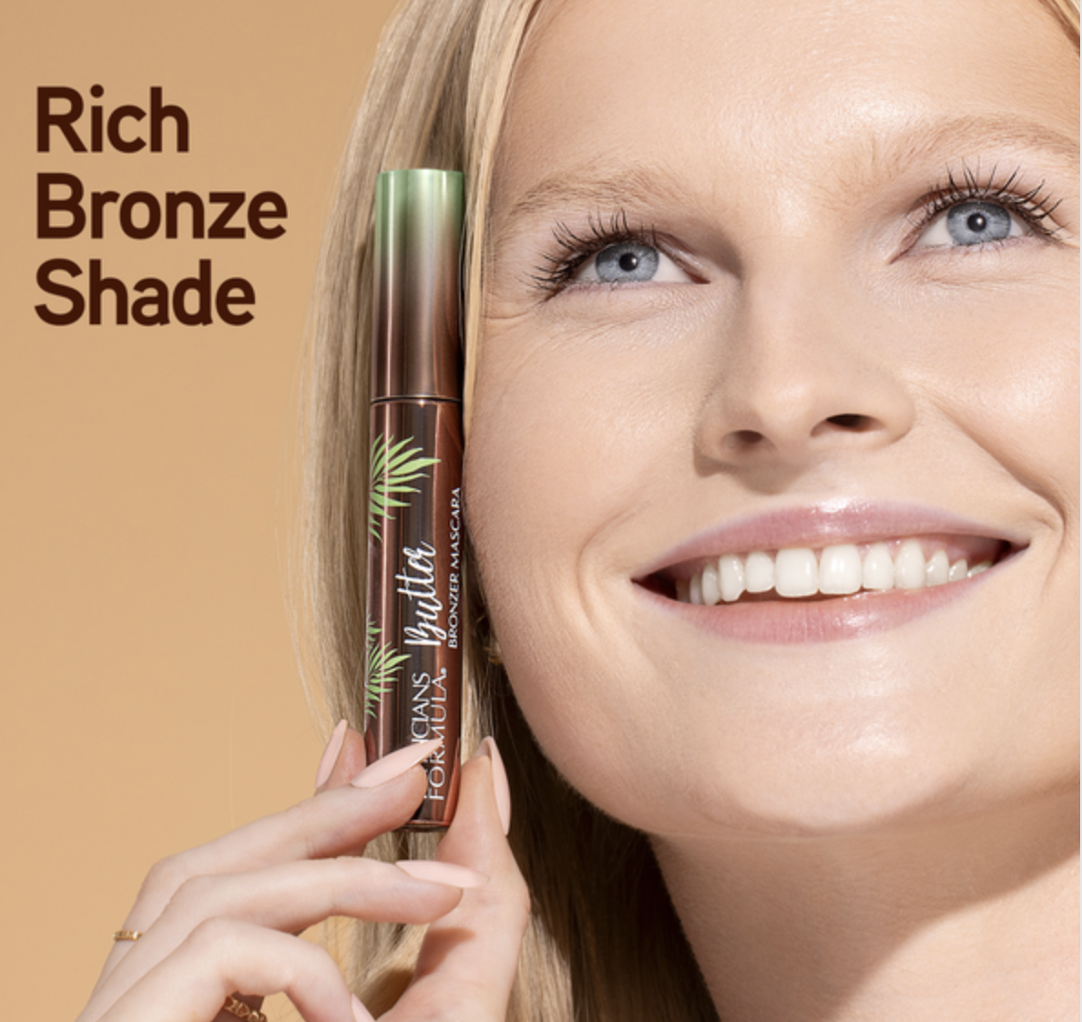 Close-up of a smiling woman with blue eyes holding a mascara tube to her face. The mascara tube is brown with green accents and labeled 'L'Oreal Paris.' Text on the left side reads 'Rich Bronze Shade.' Commercial Photographer Los Angeles.