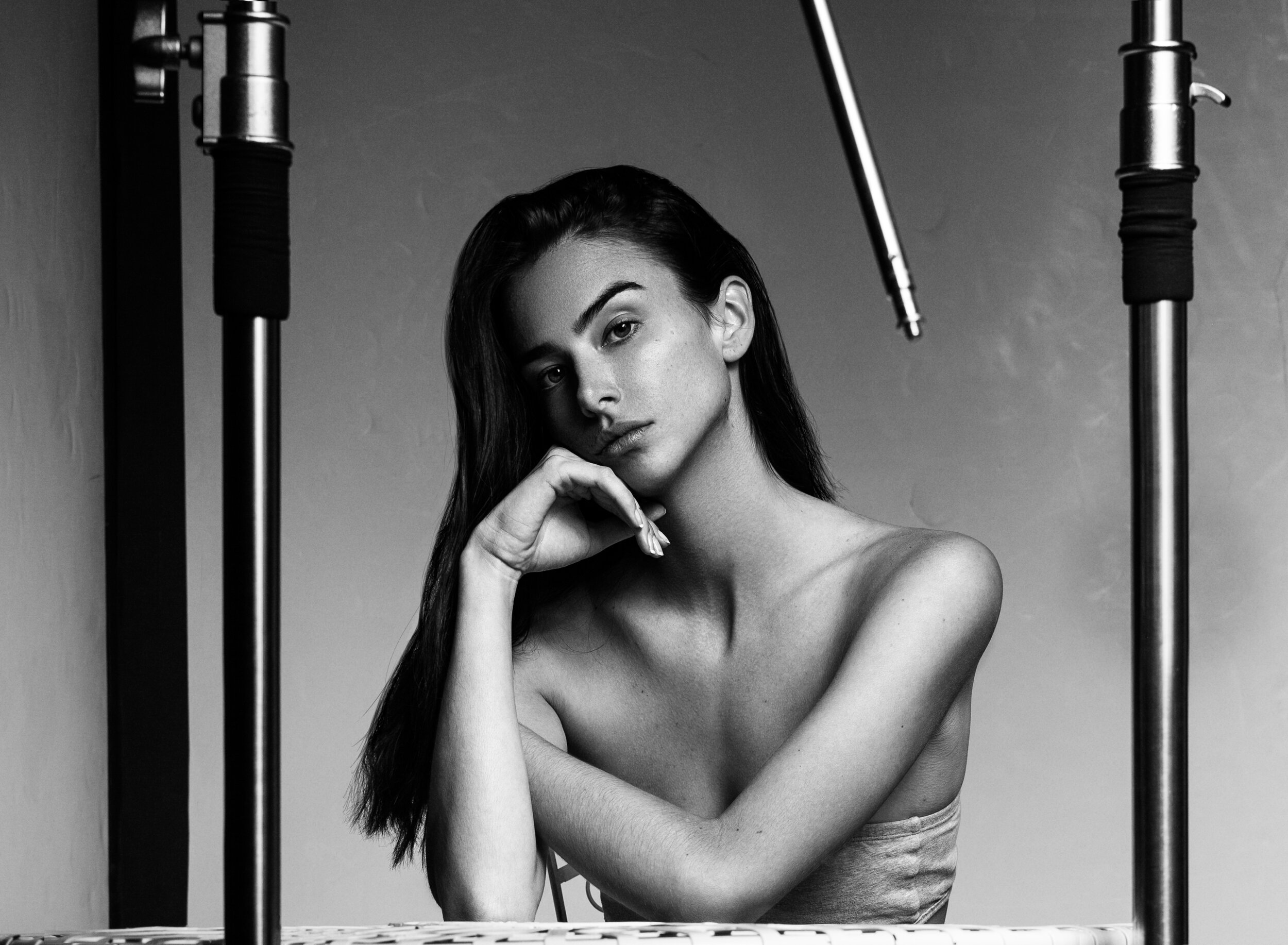 A black-and-white portrait of a woman with long dark hair, resting her chin on her hand, surrounded by studio lighting equipment.