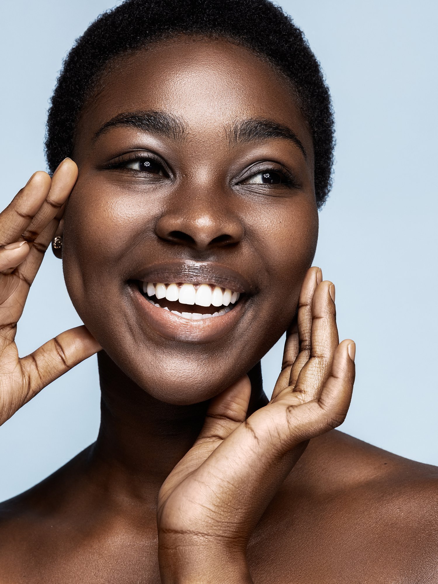 Close-up of a smiling Black woman with short natural hair, touching her face with both hands, against a light blue background. Beauty Photographer Los Angeles.