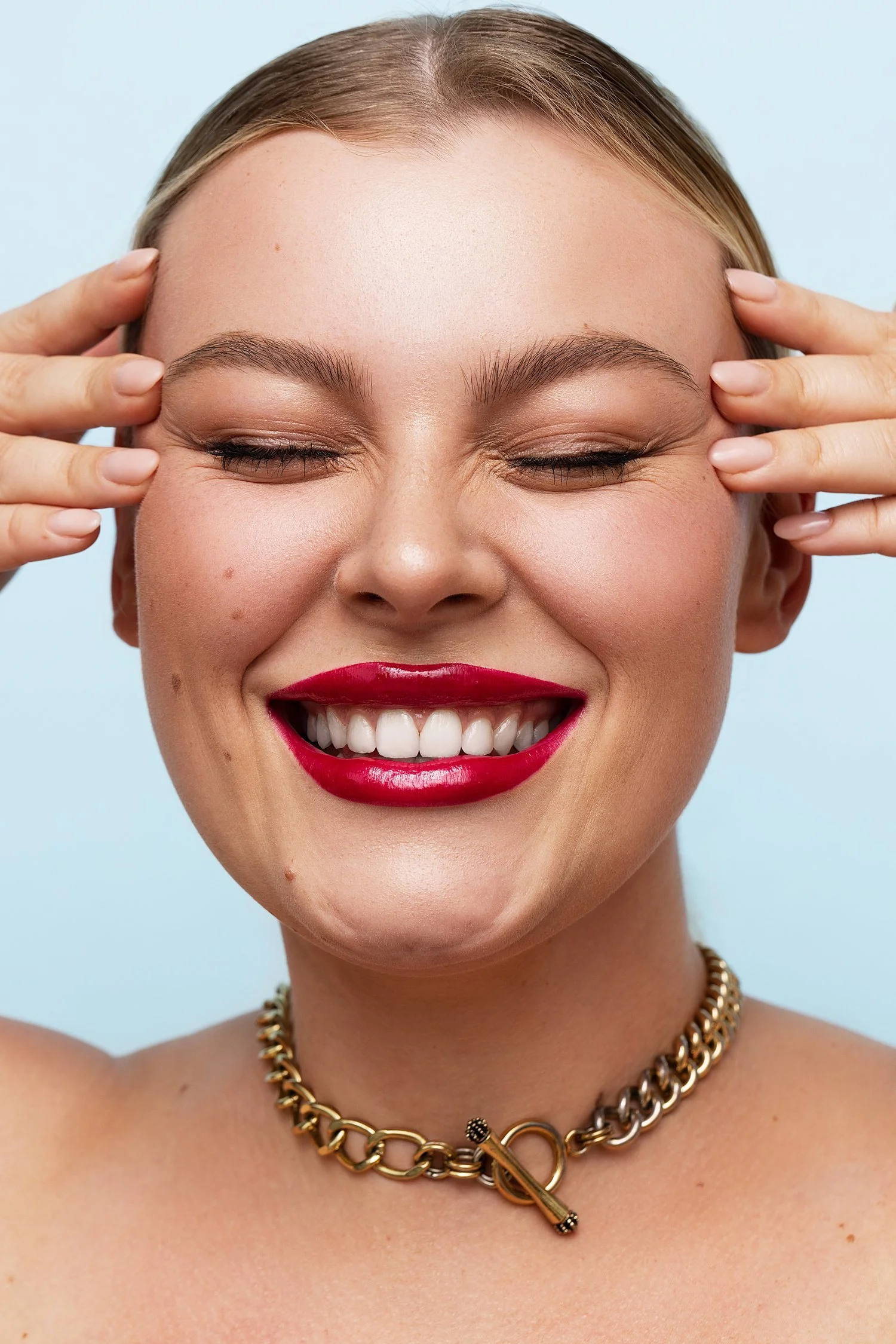 A woman with blonde hair smiling with eyes closed, wearing red lipstick, a gold chain necklace, and a gold latch choker, with hands on her temples, against a light blue background. Beauty Photographer Los Angeles.