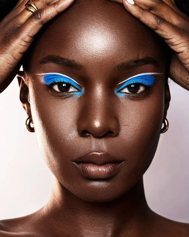 Close-up portrait of a woman with dark skin and bold white and blue eye makeup, holding her head with both hands, wearing small hoop earrings. Beauty Photographer Los Angeles.