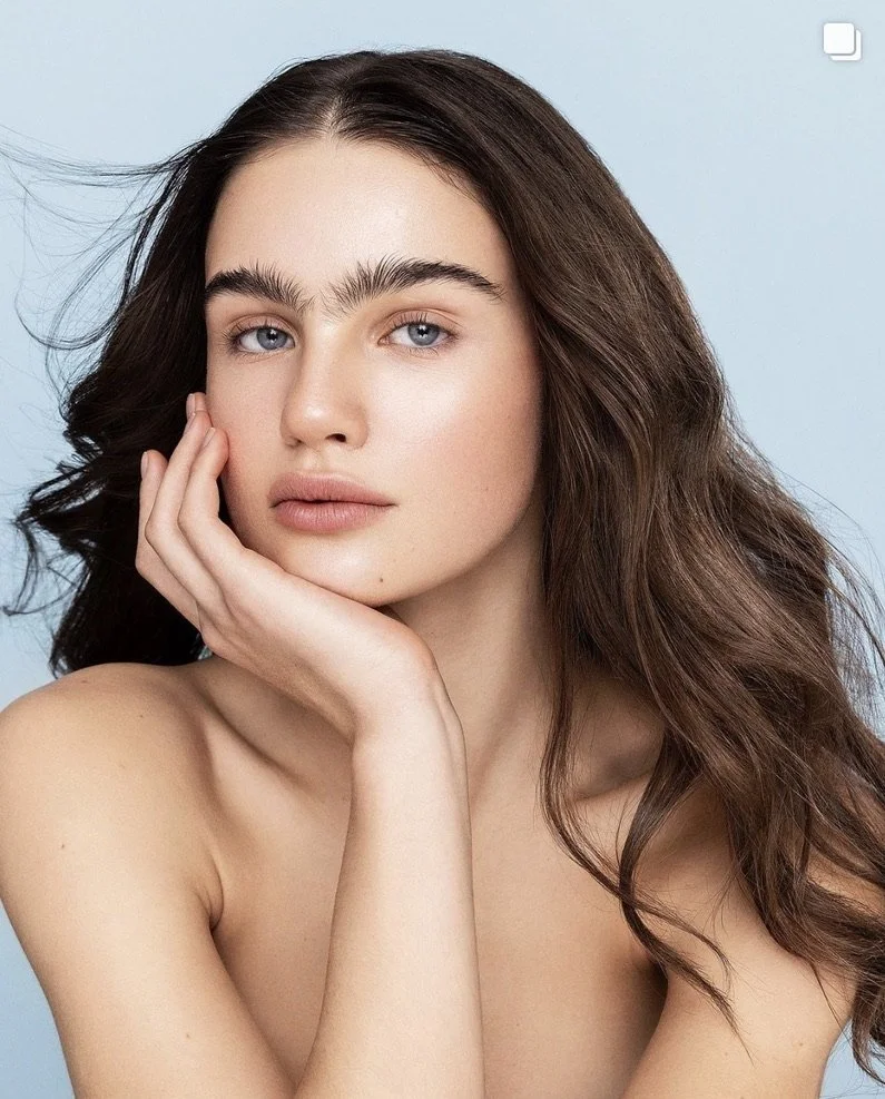 Close-up portrait of a young woman with long, wavy brown hair, fair skin, and blue eyes, resting her face on her hand with a calm expression, against a light blue background. Beauty Photographer Los Angeles.