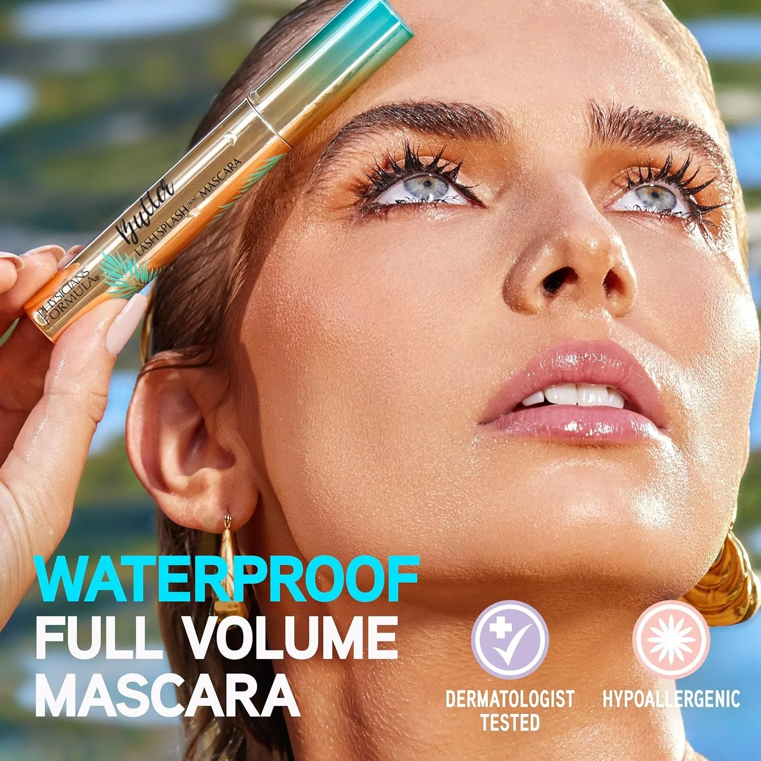 Close-up of a woman with wet hair, holding a tube of waterproof full volume mascara near her temple. She has bright blue eyes, long lashes, and is outdoors with blurred background. Text overlay says 'Waterproof Full Volume Mascara' with icons indicat