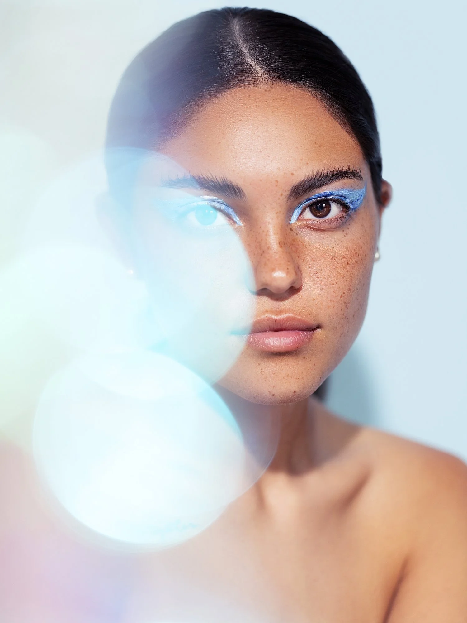 Close-up portrait of a young woman with dark hair, natural freckles, and blue eye makeup, looking directly at the camera against a light background with bokeh lights. Beauty Photographer Los Angeles.