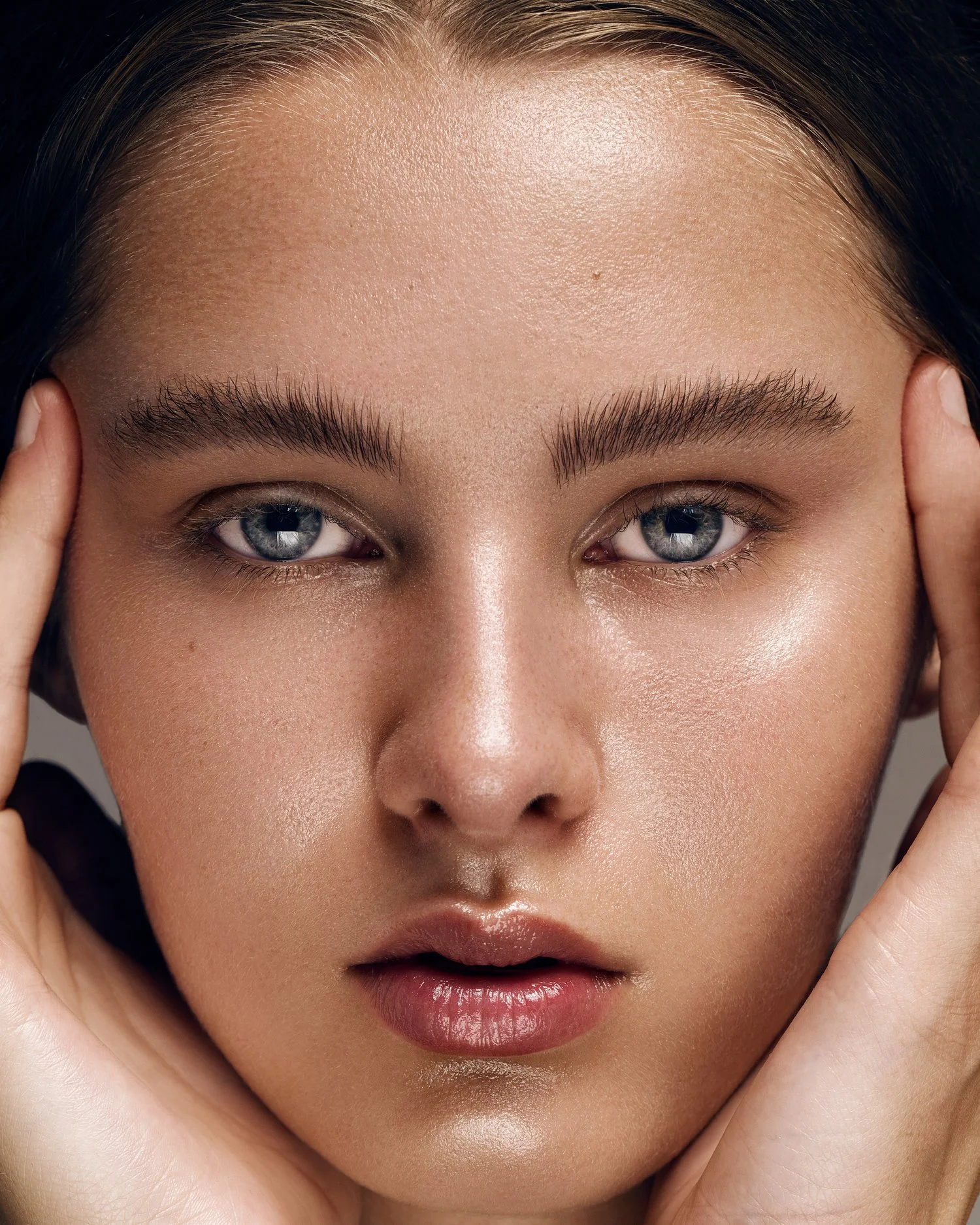 Close-up of a woman's face with clear skin, blue eyes, and dark hair, holding her face with her hands. Beauty Photographer Los Angeles.