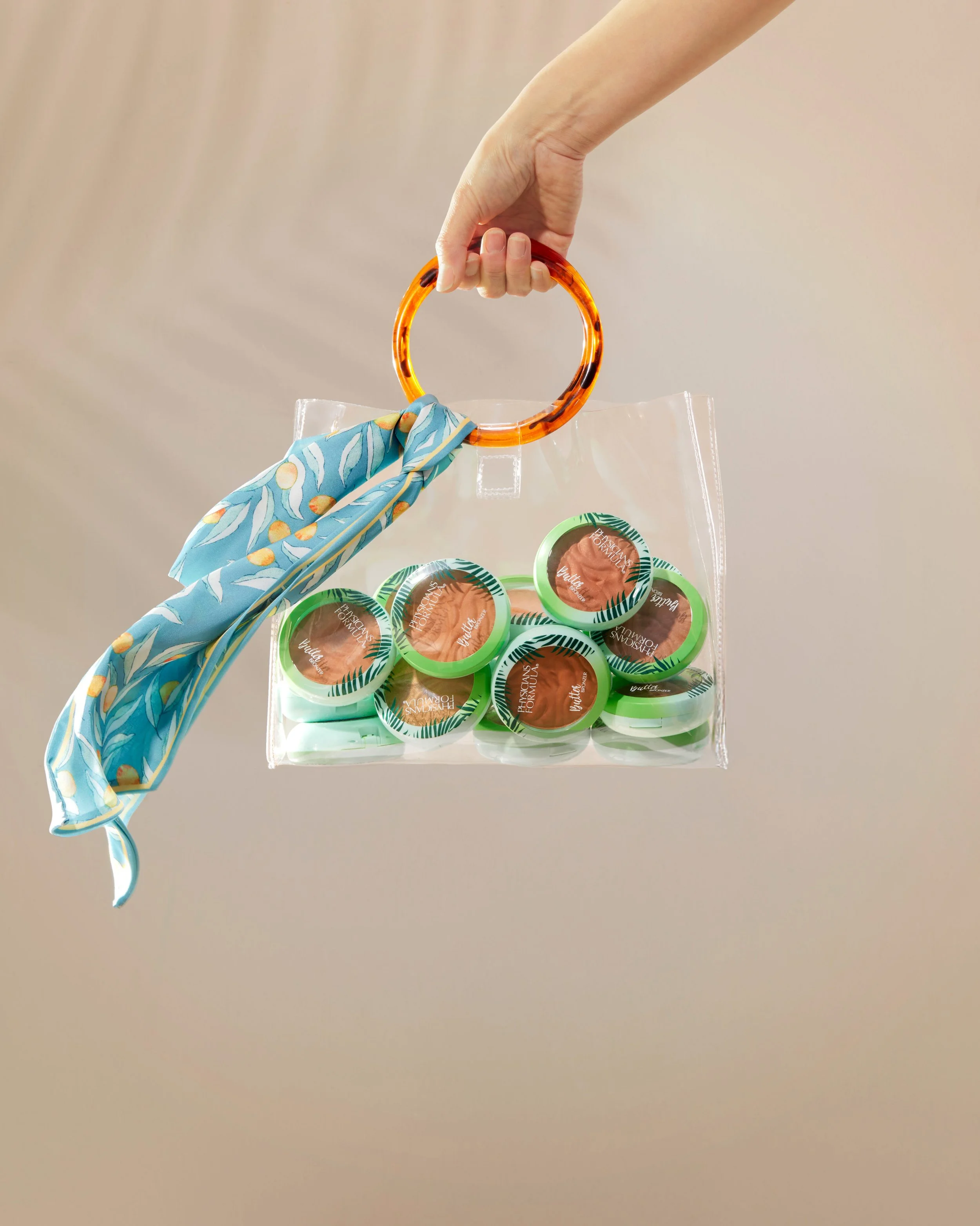 A hand holding a clear plastic bag filled with small containers of a brown substance, with a blue scarf tied to an orange tortoise shell patterned ring attached to the bag. The background is a plain beige wall. Commercial Photographer Los Angeles.