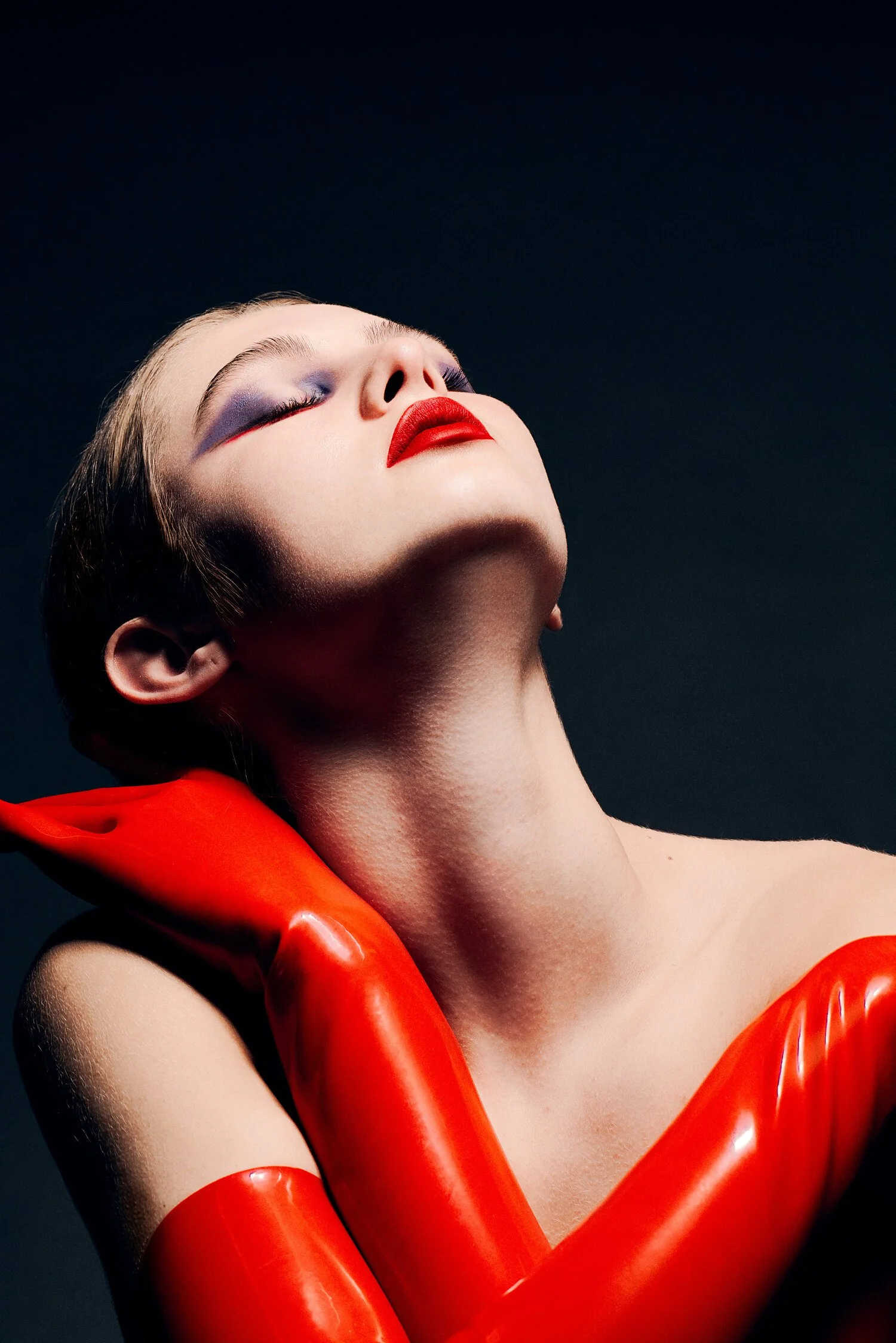 A woman with bold makeup, wearing red gloves, and a red lipstick. Her face is tilted back with eyes closed, against a dark background. Beauty Photographer Los Angeles.