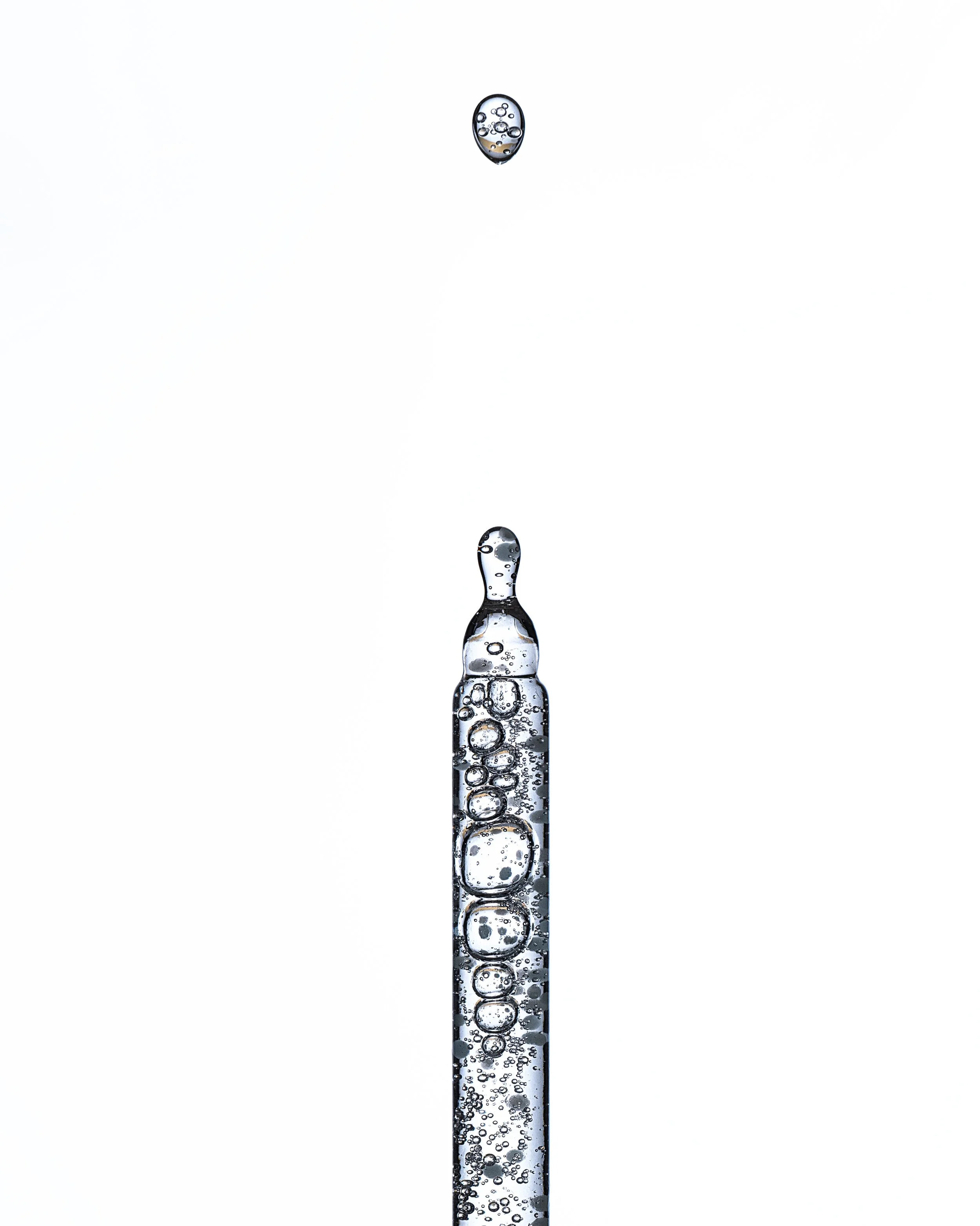 A close-up of a syringe filled with a clear liquid and bubbles, with a drop of liquid falling from the tip against a plain white background. Product photography Los Angeles.