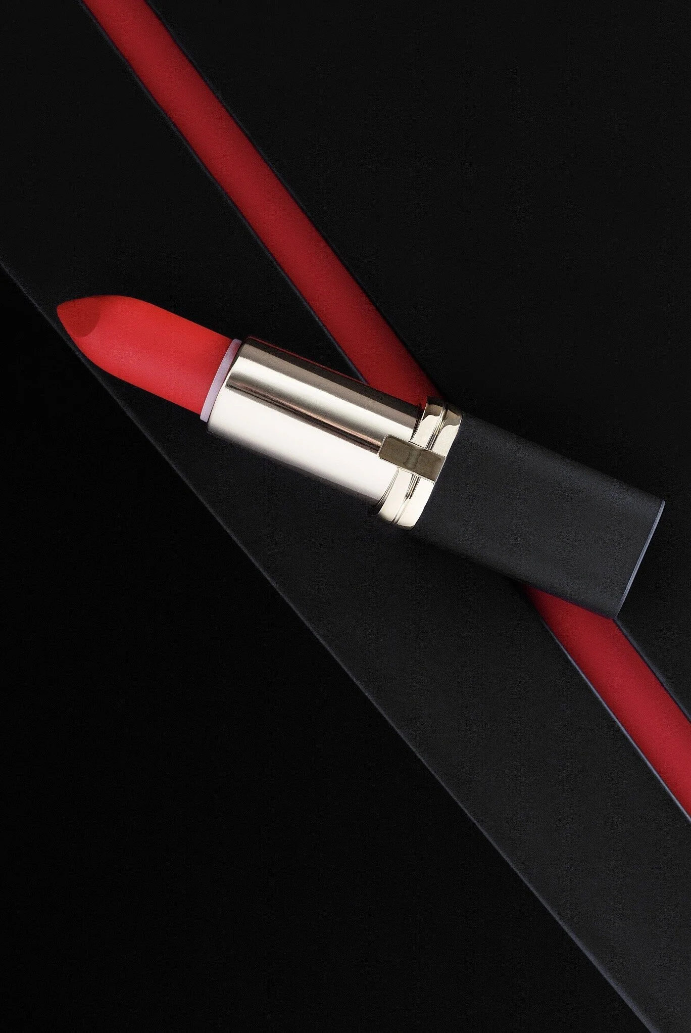 A close-up photograph of a red lipstick with black and gold casing on a black background with red accents. Product photography Los Angeles.