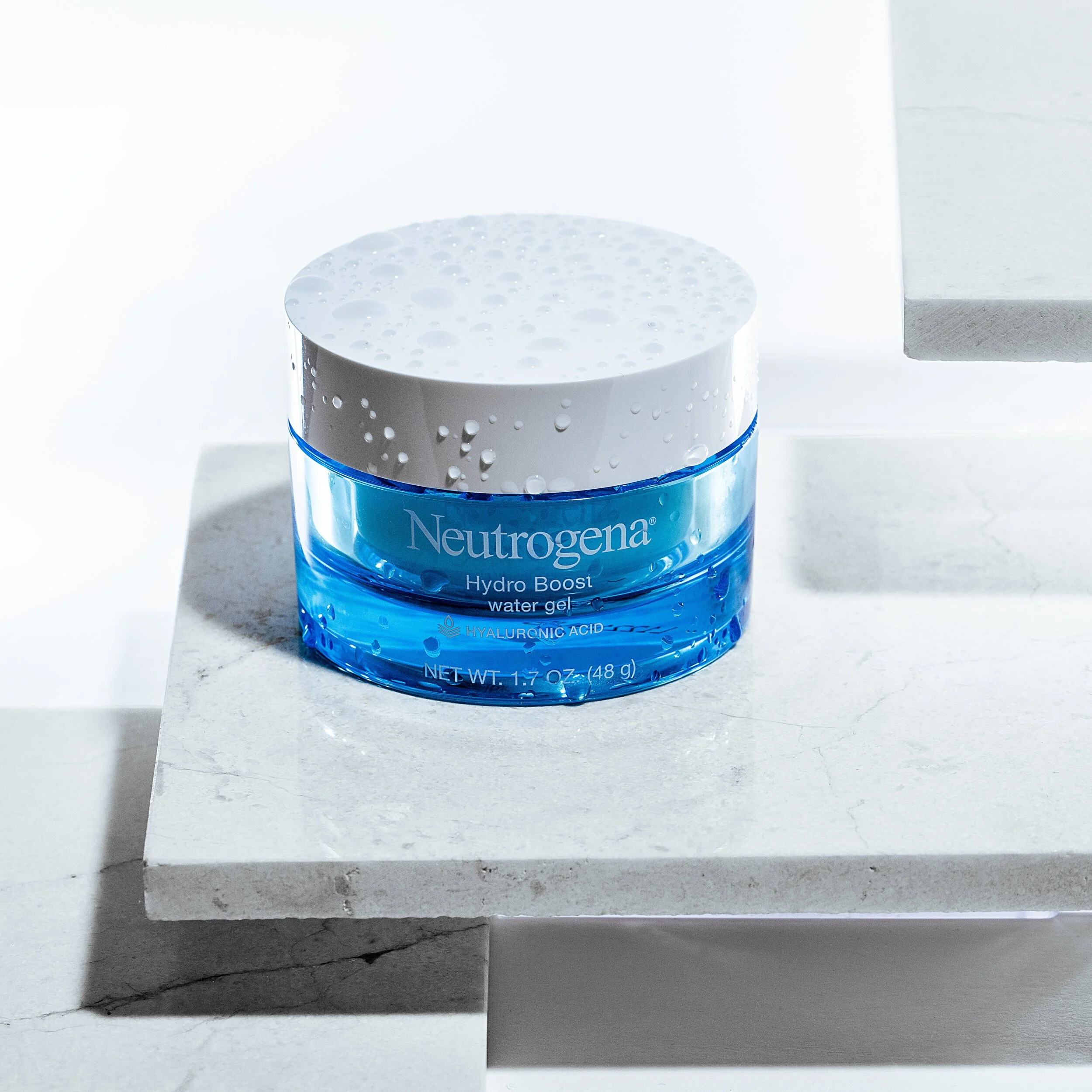 A jar of Neutrogena Hydro Boost water gel with hyaluronic acid sitting on a white marble surface, with water droplets on the jar and a white background.