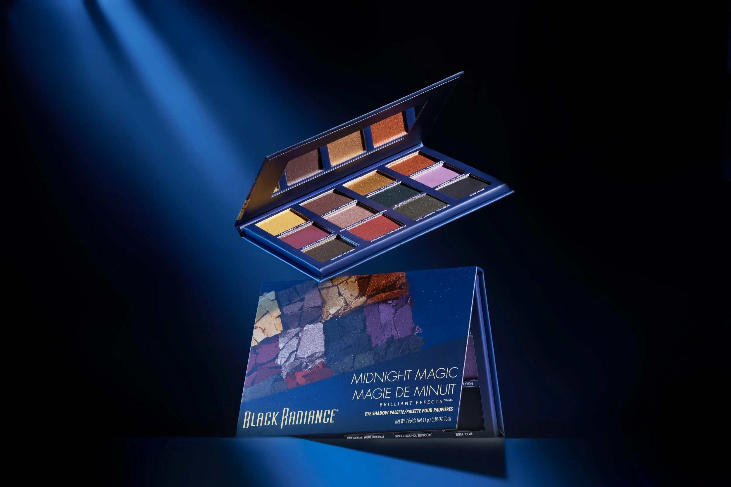 Open eyeshadow palette with 12 colorful shades, titled 'Midnight Magic' from Black Radiance, with a matching box underneath against a dark background with blue lighting. Product photography Los Angeles.