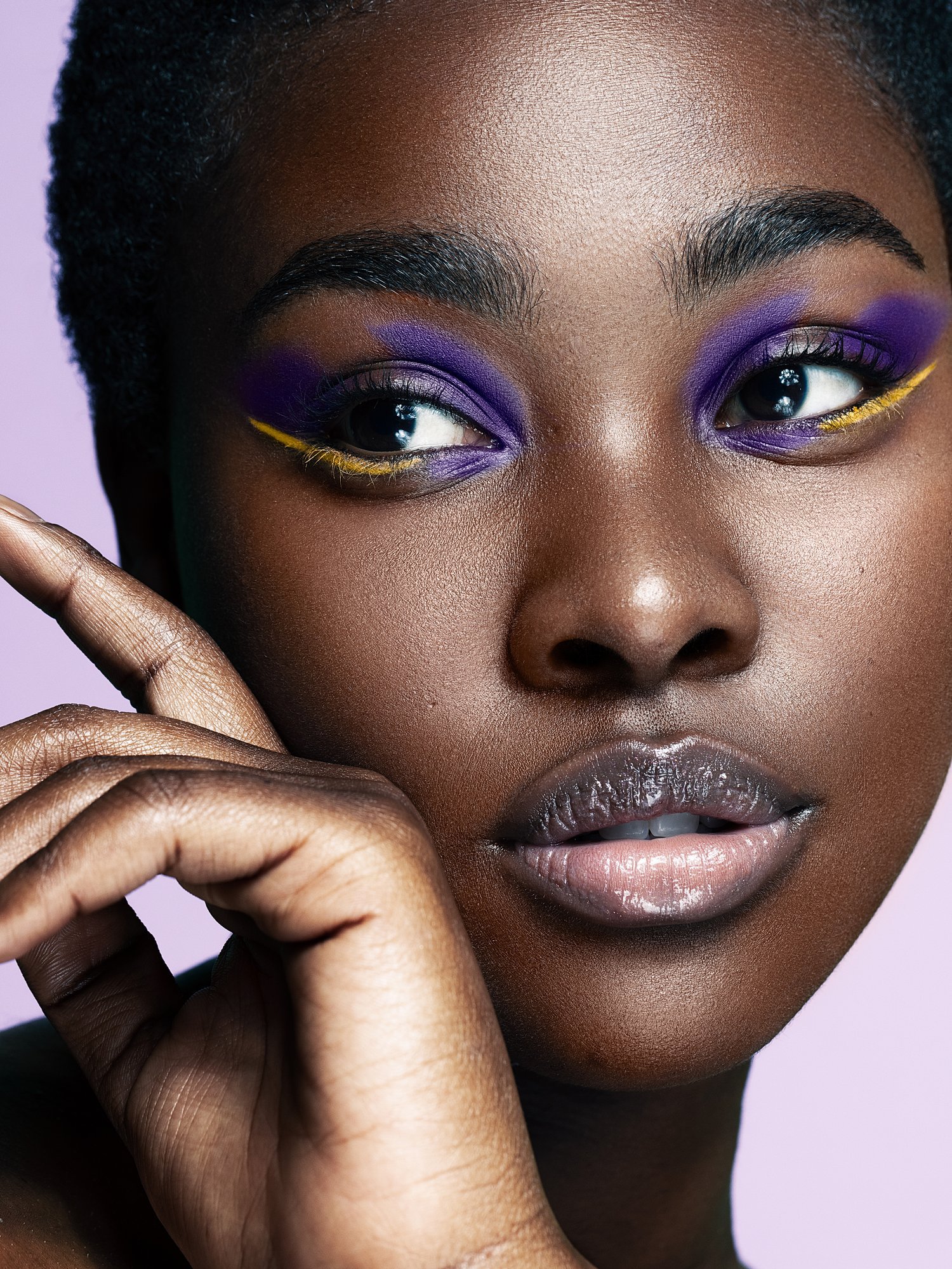Close-up of a woman's face with vibrant purple and yellow eye makeup, glossy lips, and smooth skin, with a light purple background. Beauty Photographer Los Angeles.