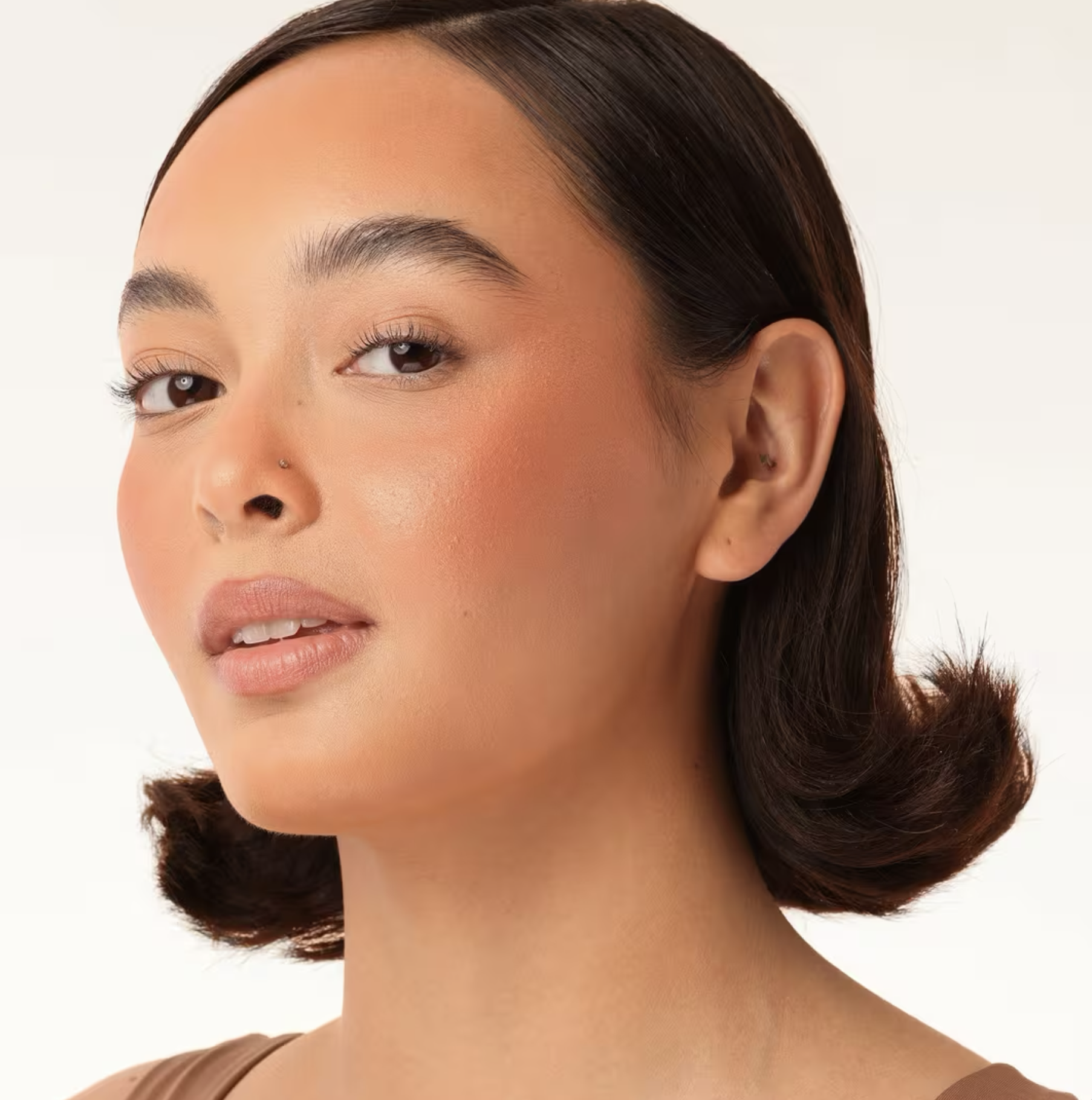 Close-up of a woman with short dark hair, natural makeup, and a small nose piercing, looking confidently at the camera against a plain background.