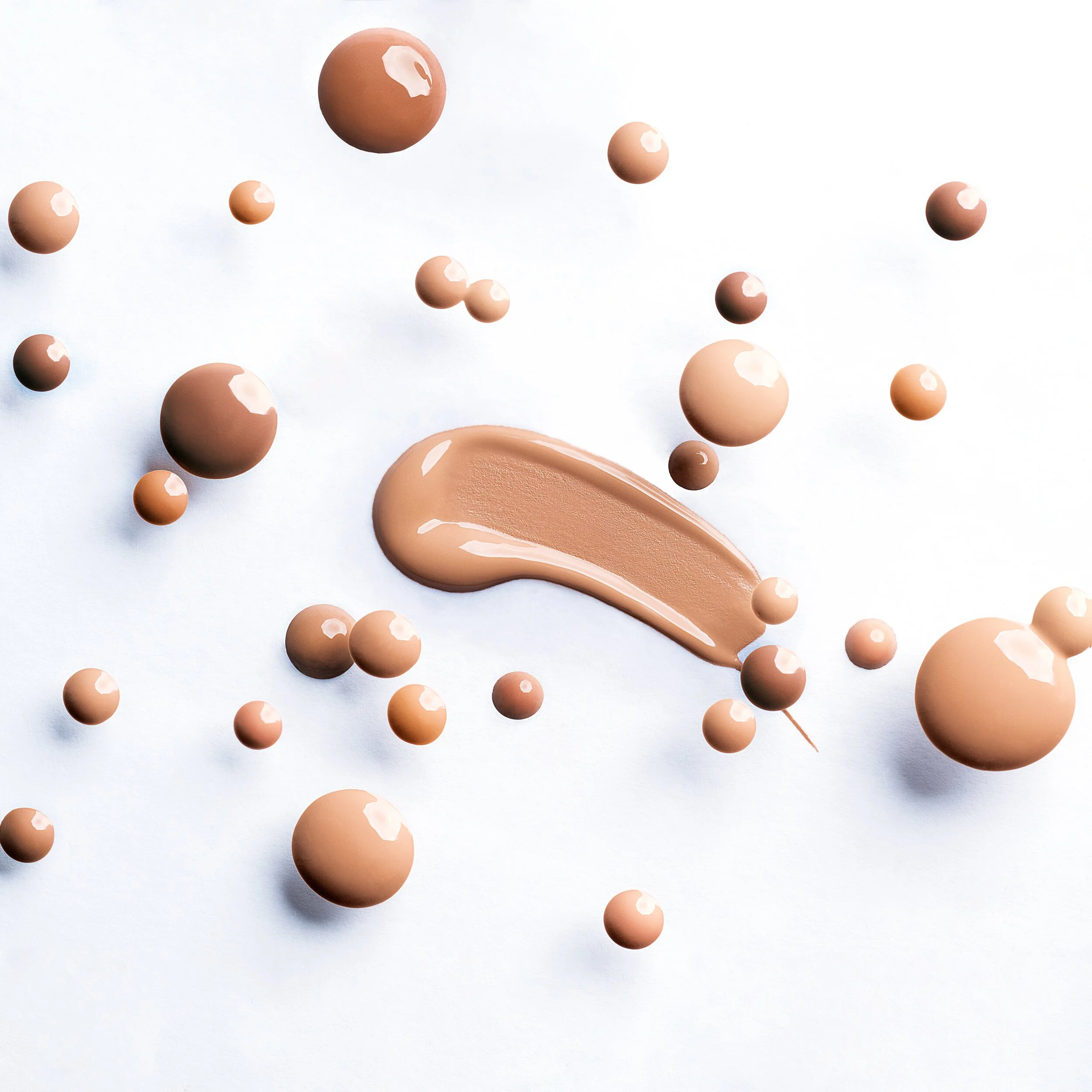 Cream foundation spread with scattered spherical drops of the same beige shade on a white background