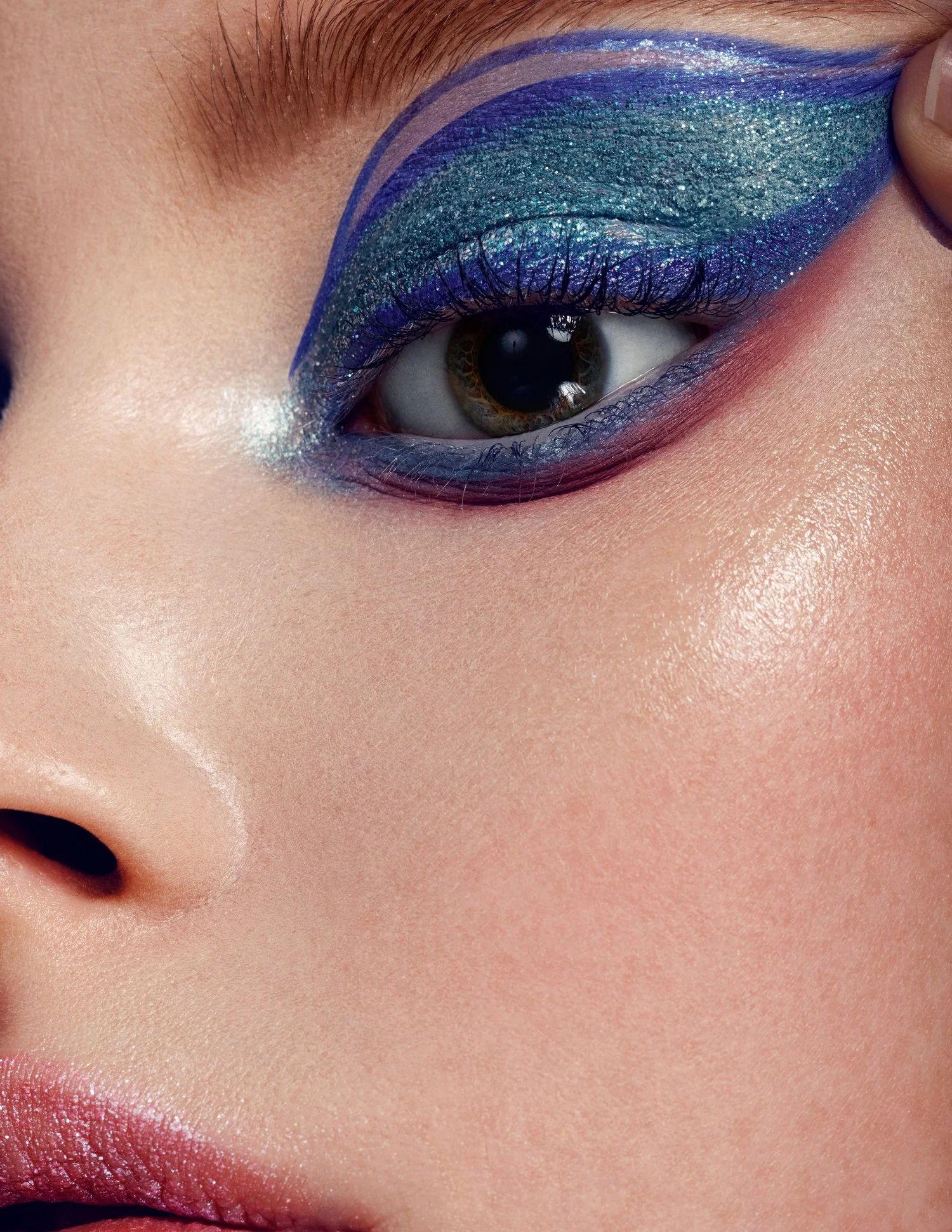 Close-up of a person's eye with elaborate blue and green glitter eyeshadow, accented with purple eyeliner and white shimmer, with visible skin texture and pink lips. Beauty Photographer Los Angeles.