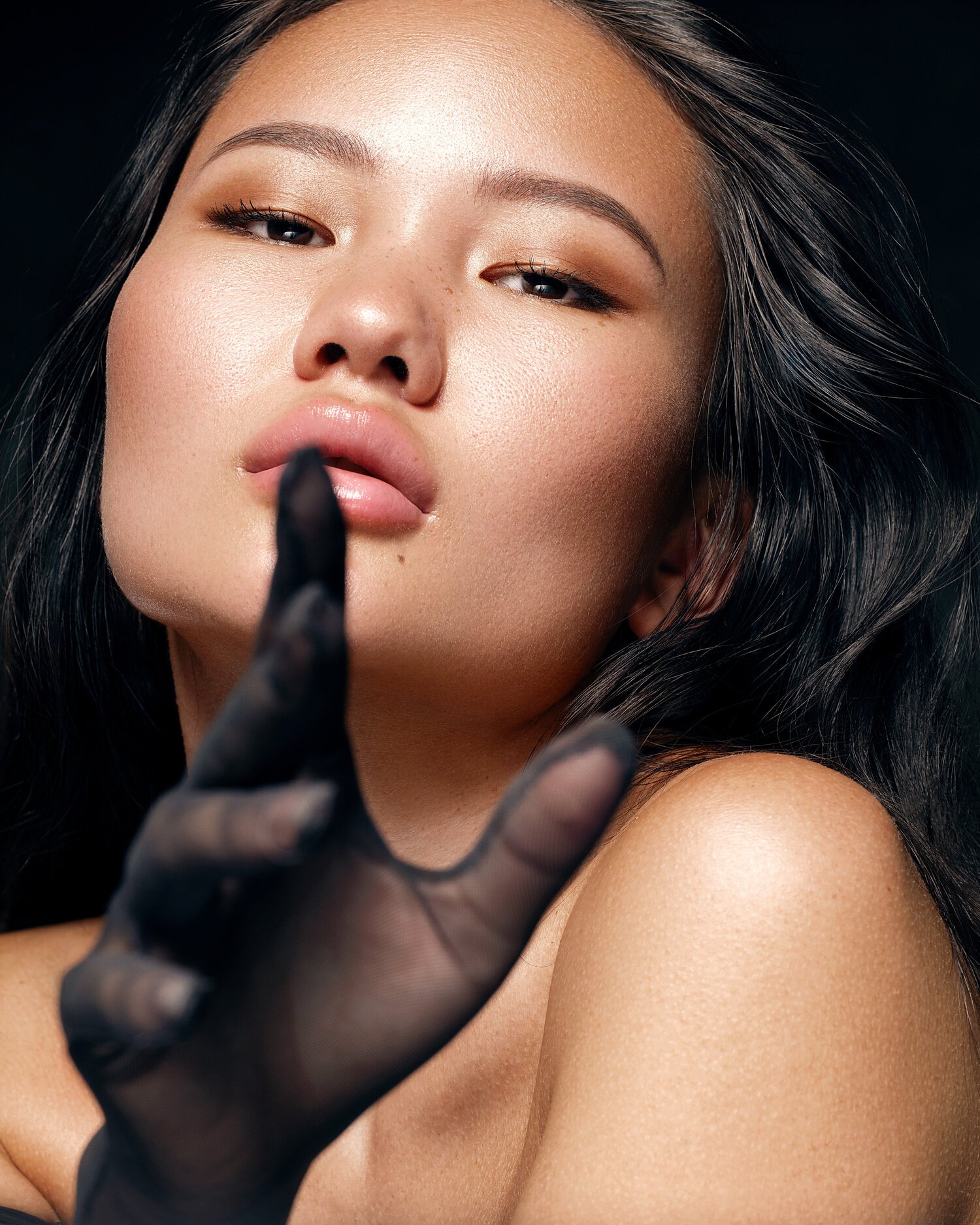 A close-up of a woman with dark hair, wearing subtle makeup, with her finger, in a black glove, held up in front of her lips, making a shushing gesture. Beauty Photographer Los Angeles.