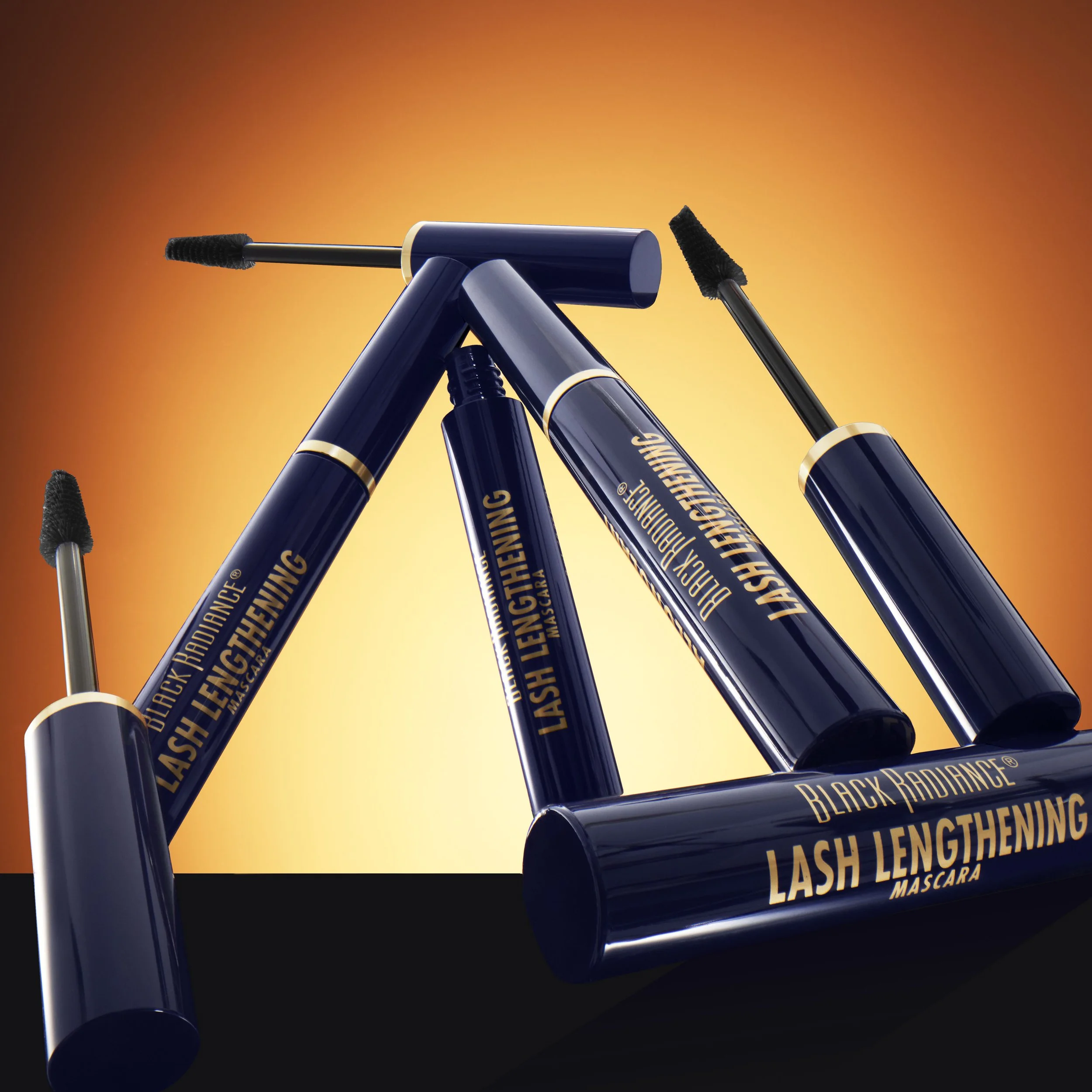 Multiple tubes of Black Radiance lash lengthening mascara arranged on a dark surface with an orange gradient background. Product photography Los Angeles.