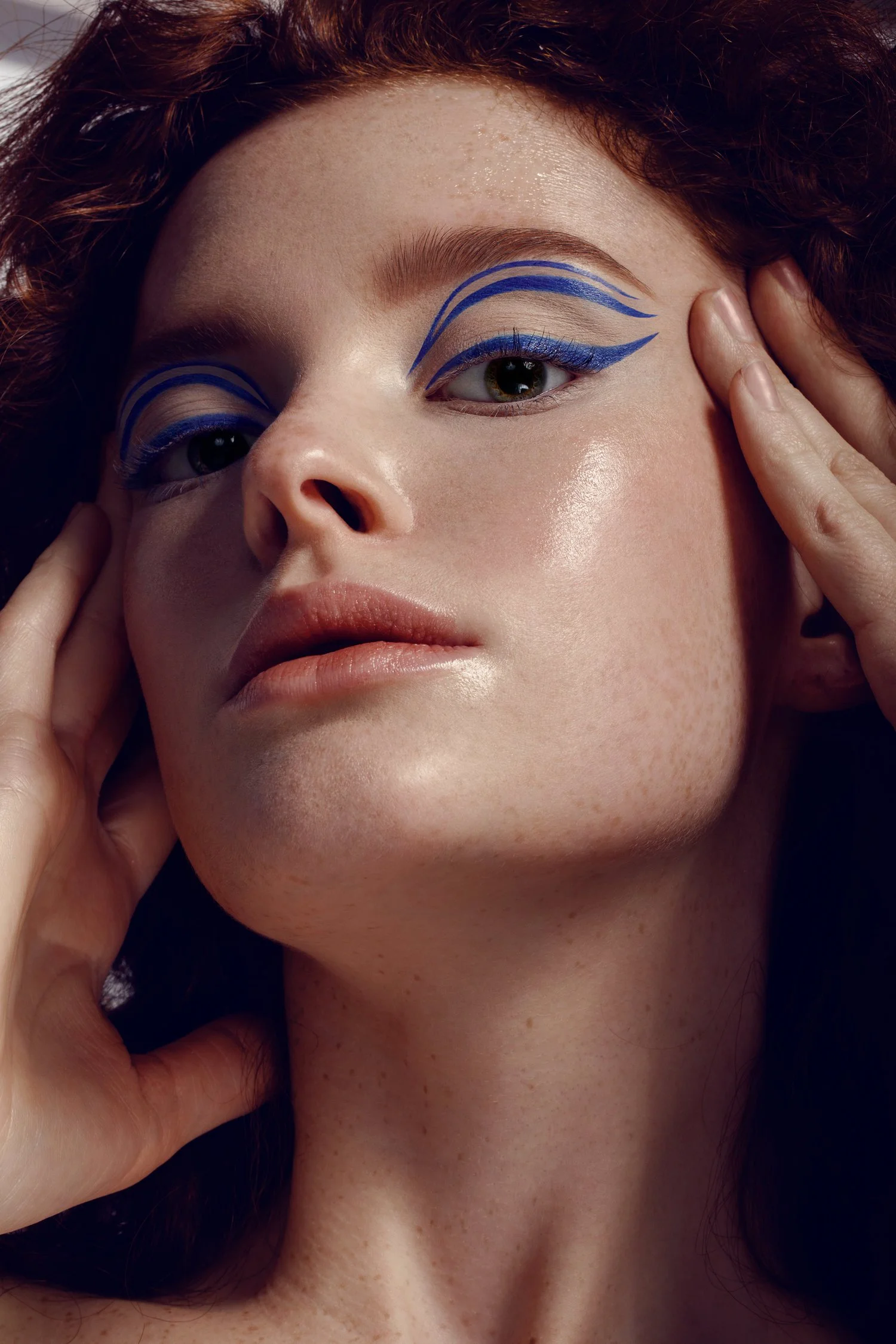 Close-up of a woman with blue graphic eyeliner, touching her face with both hands. Beauty Photographer Los Angeles.