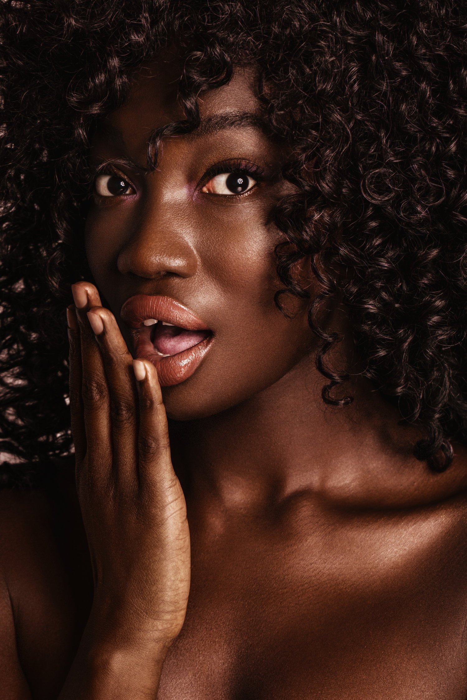 A close-up portrait of a woman with dark curly hair, expressive eyes, and lips slightly parted, touching her face with her hand. Beauty Photographer Los Angeles.