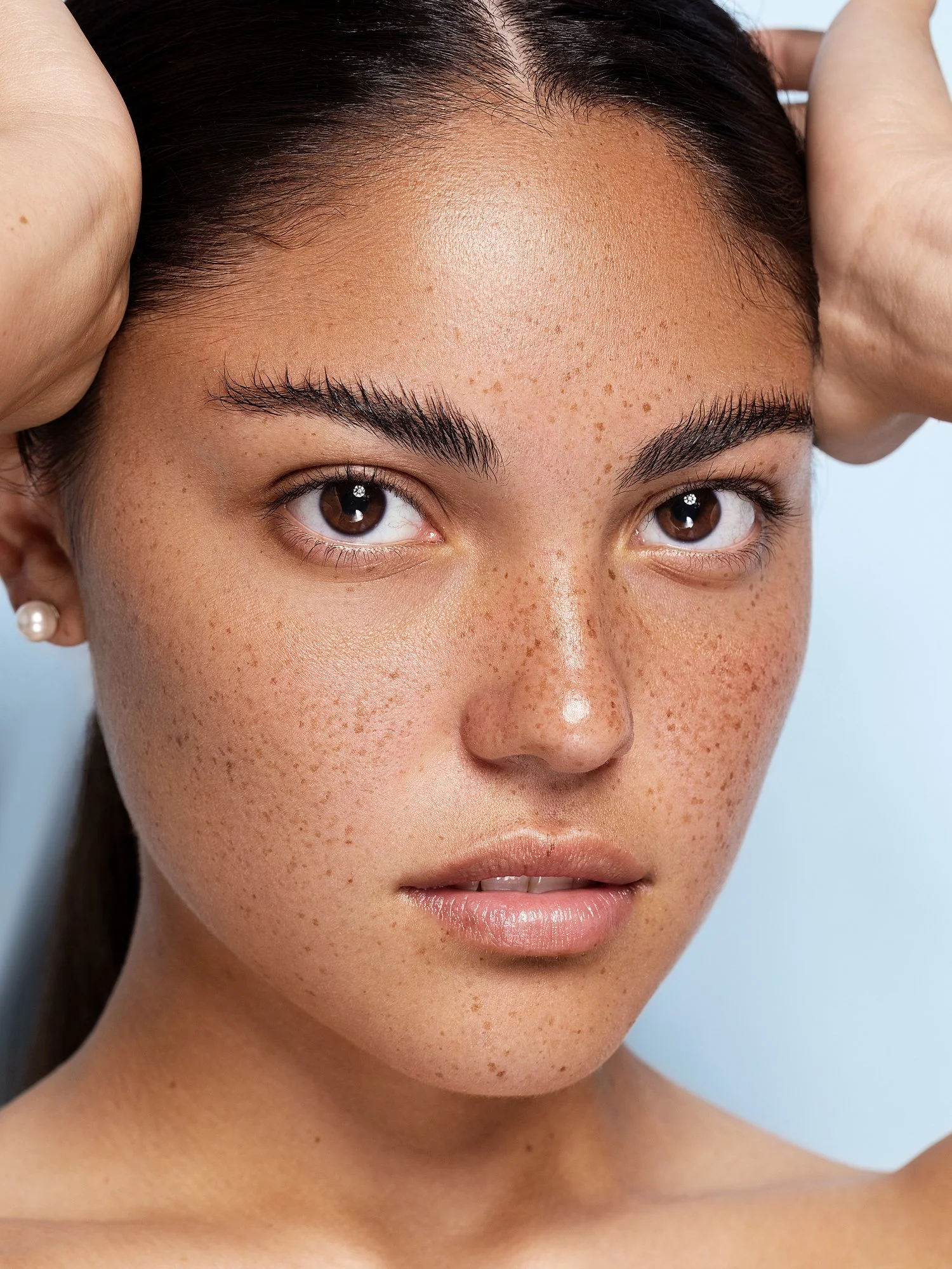 Close-up of a woman's face with brown eyes, dark hair, and visible freckles, holding her hands behind her head. Beauty Photographer Los Angeles.