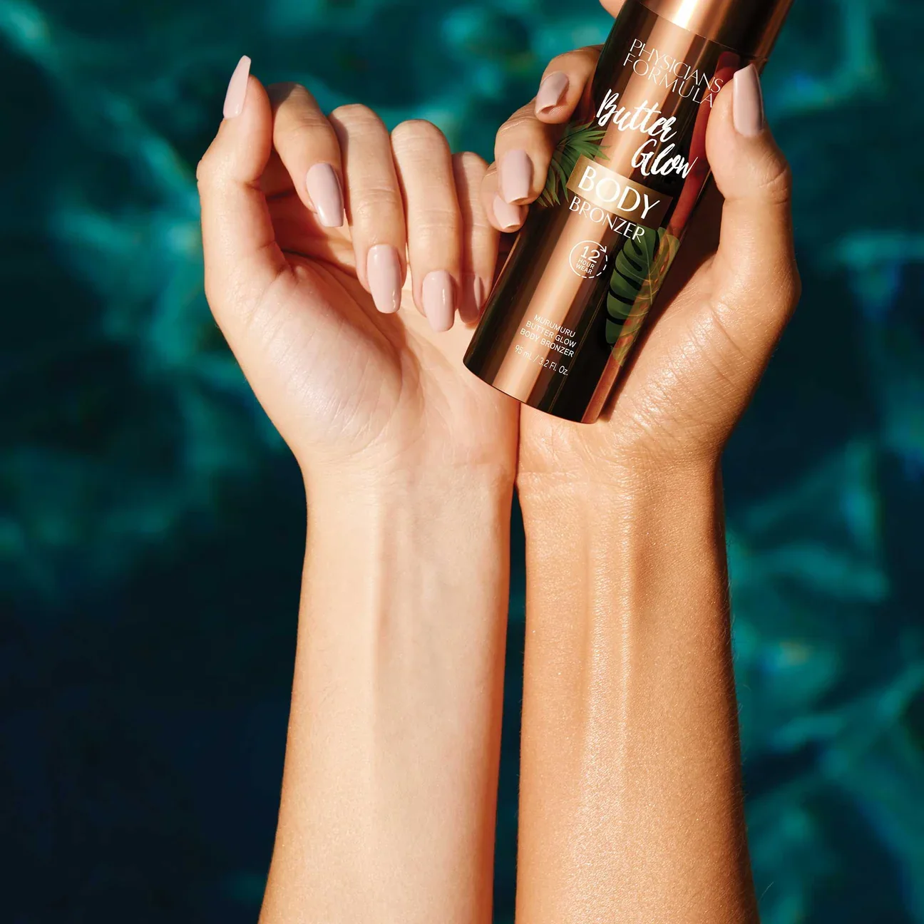 A person holds a bronze-colored bottle of Physicians Formula Butter Glow Body Bronzer with two arms, one with fair skin and the other with darker skin, against a blue and green background.