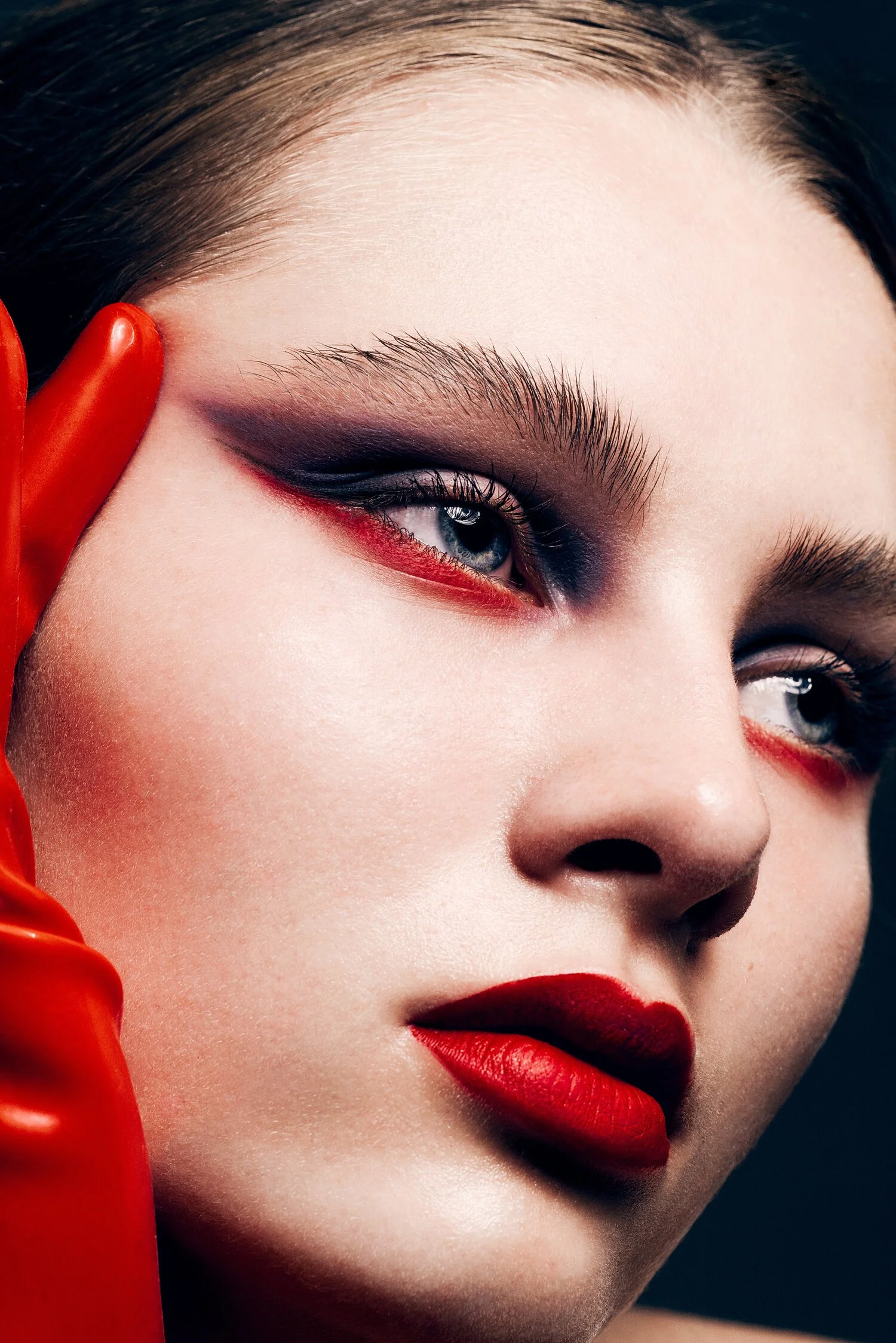 A close-up of a woman's face with bold red lipstick, striking eye makeup, and a red glove touching her face. Beauty Photographer Los Angeles.