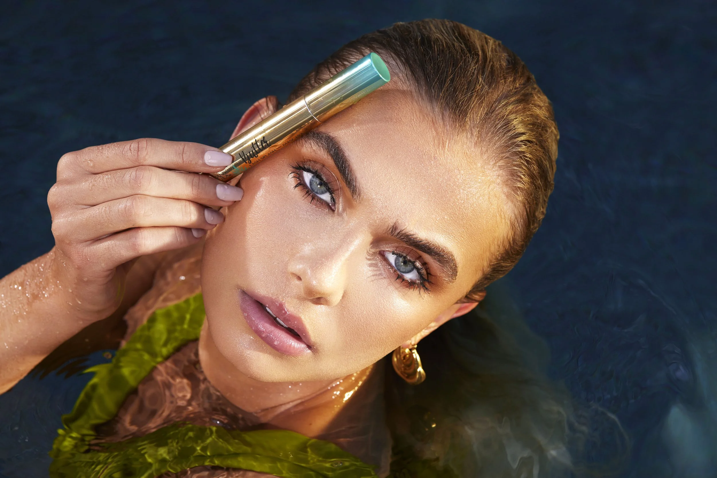 A woman with wet hair and makeup holding a gold and turquoise eyeliner stick near her eyebrow while floating in water, wearing large gold earrings and a green leafy top. Commercial Photographer Los Angeles.