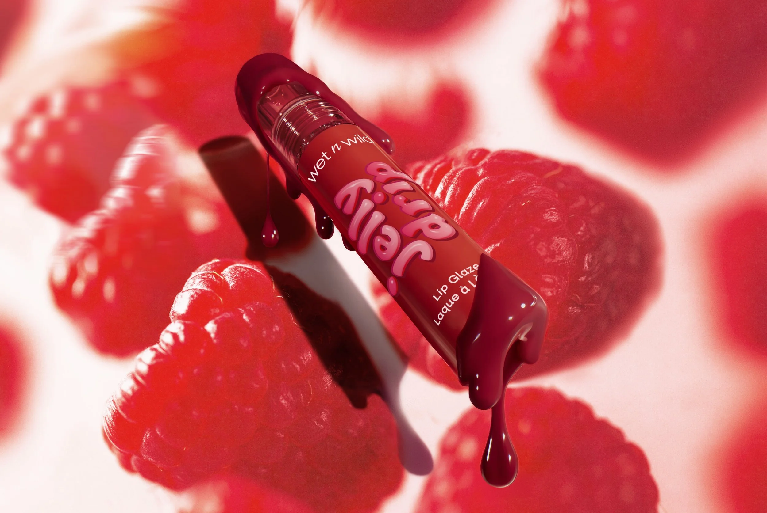 A raspberry-colored lip gloss tube labeled 'Wet n Wild Lip Gloss' is lying on a background featuring close-up raspberries. Some of the lip gloss is dripping from the tube. Product photography Los Angeles.
