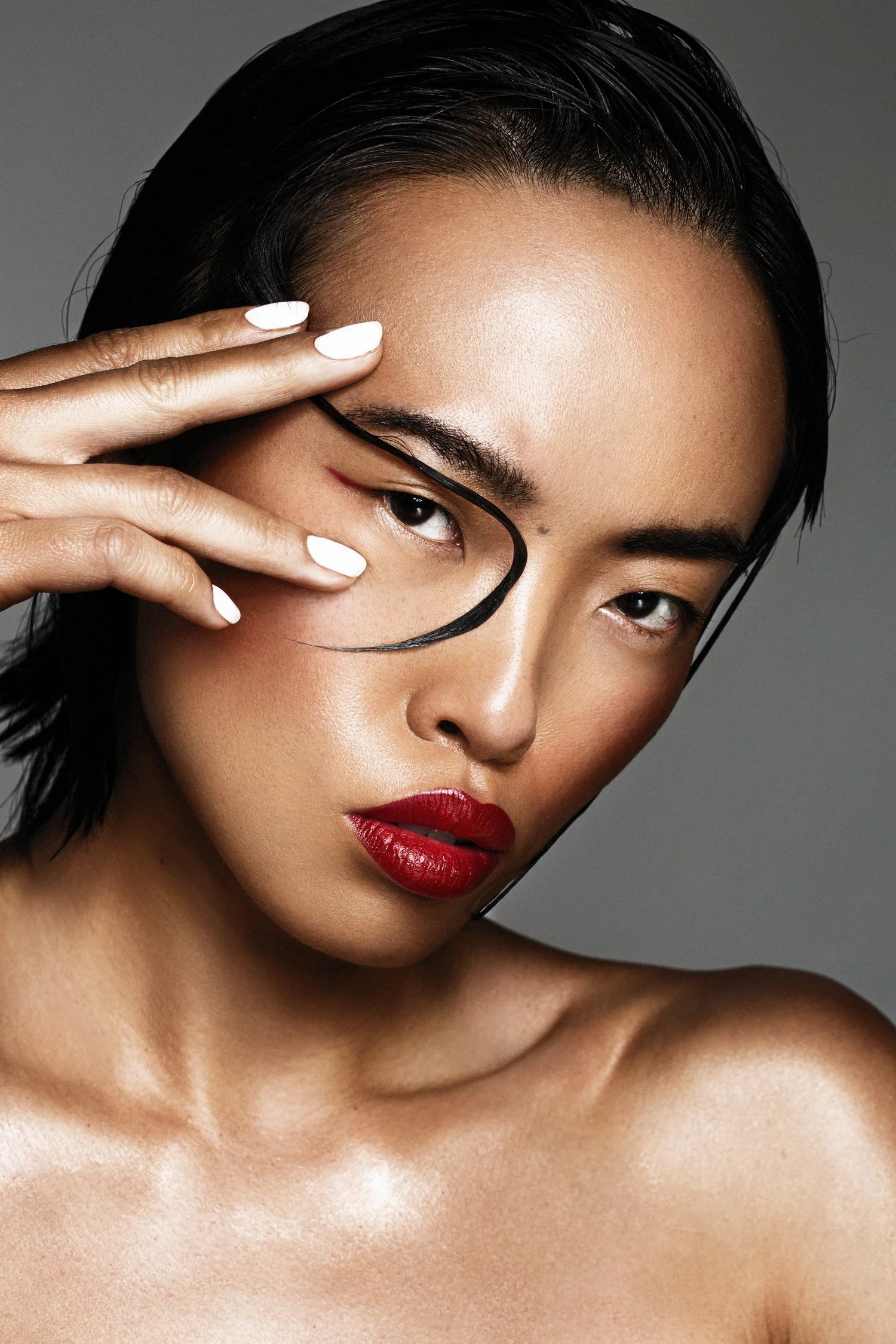 Close-up portrait of a woman with wet black hair, red lipstick, and white nail polish, posing with her hand near her face. Beauty Photographer Los Angeles.