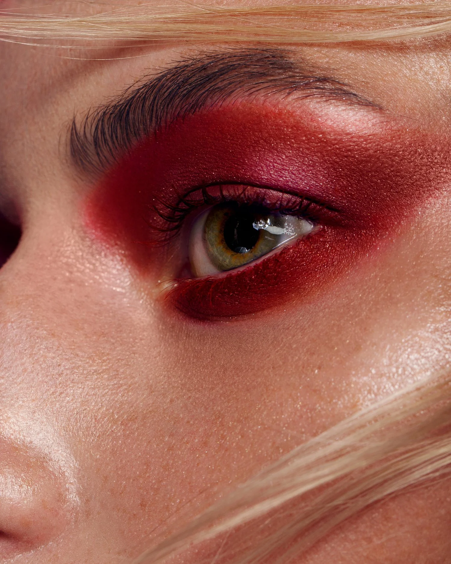 Close-up of a person's eye with bold red eyeshadow and eyeliner, with visible skin texture and light-colored hair at the bottom. Beauty Photographer Los Angeles.